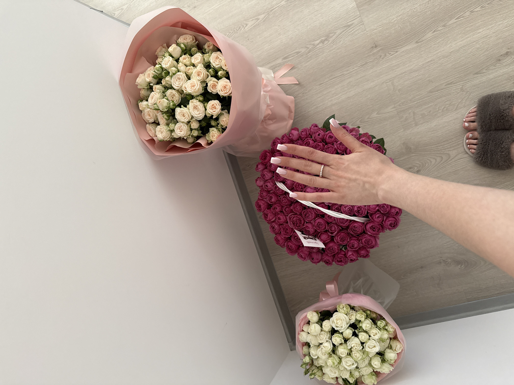 In an indoor setting, likely a room with light-colored walls and a wooden floor, three bouquets of roses are arranged. One bouquet, comprised of dark pink roses, is prominently displayed in the center foreground. A hand, adorned with a ring and manicured nails, is positioned over this bouquet. To the left, a smaller bouquet of white roses is situated, partially wrapped in pink paper. To the right, another bouquet of cream-colored roses, also wrapped in pink paper, leans against the wall. The overall impression is one of a staged photograph, possibly for a floral arrangement or a personal collection. There are no other visible individuals or specific environmental details that would indicate a particular location within a city.