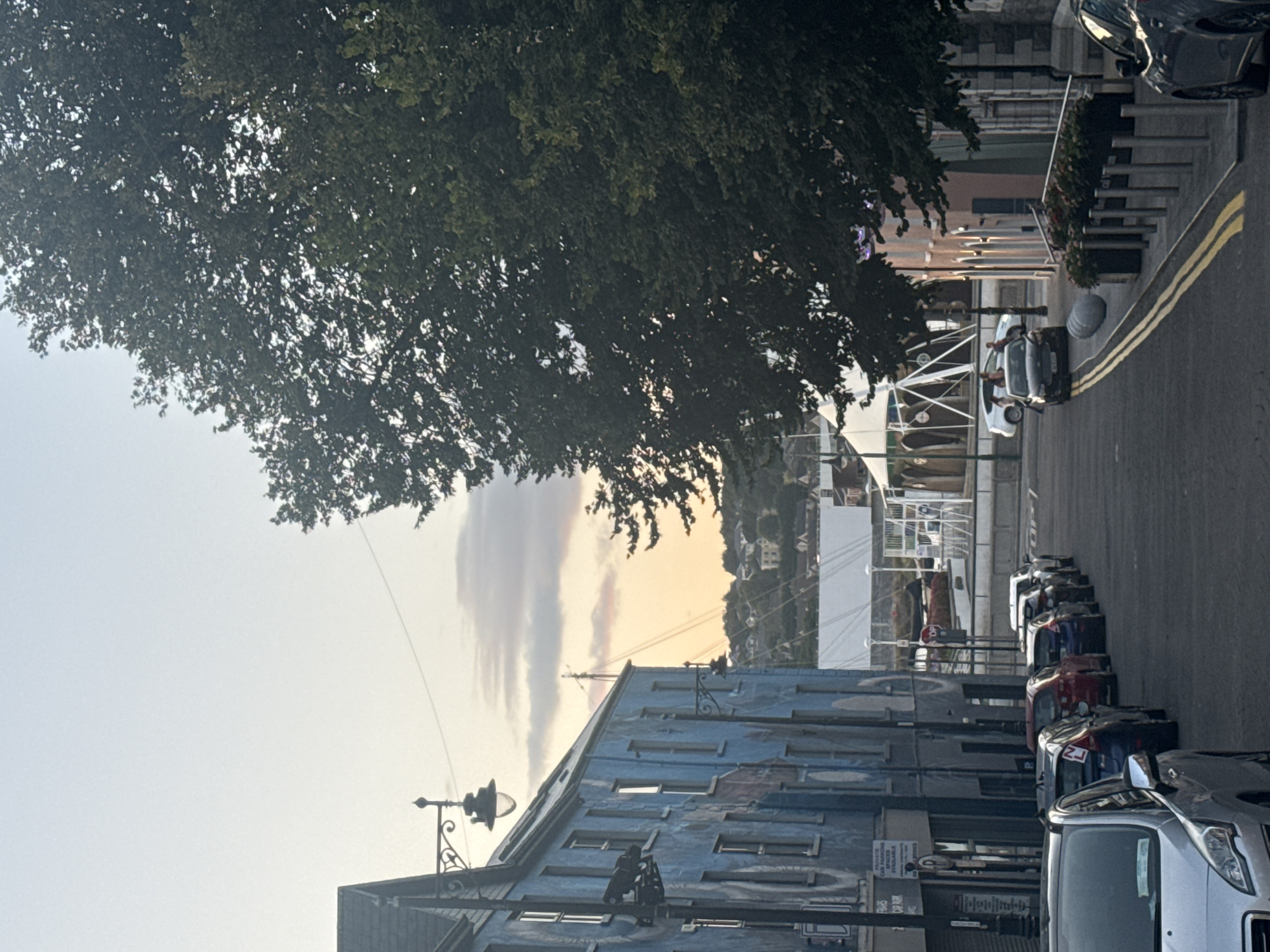The image depicts a street scene in Waterford, Ireland, with a prominent blue building on the left adorned with a mural. Several cars are parked along the street, and the scene is illuminated by the soft light of dusk or dawn. In the background, there are white tents or marquees, suggesting an event or market. The sky displays hues of orange and pink, indicating either sunrise or sunset. The street is paved, and there are lampposts and trees lining the sidewalks. The overall atmosphere is calm and quiet, with no visible human activity apart from individuals walking near the white structures in the background.