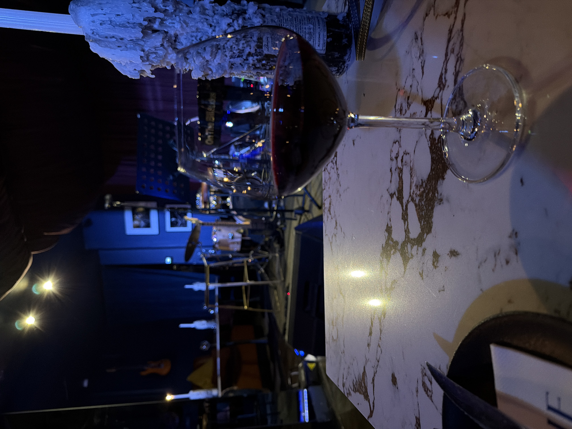 The image shows a glass of red wine on a marble-like tabletop. Behind the wine glass is a stage setting with musical instruments like a drum set and guitars, illuminated by stage lights. There's a stand holding what appears to be sheet music. To the left of the wine glass is a partially visible object covered in what resembles white wax. The image suggests a live music venue or bar, potentially in Cape Town, South Africa, though no definitive city landmarks are visible in the frame to confirm this location.