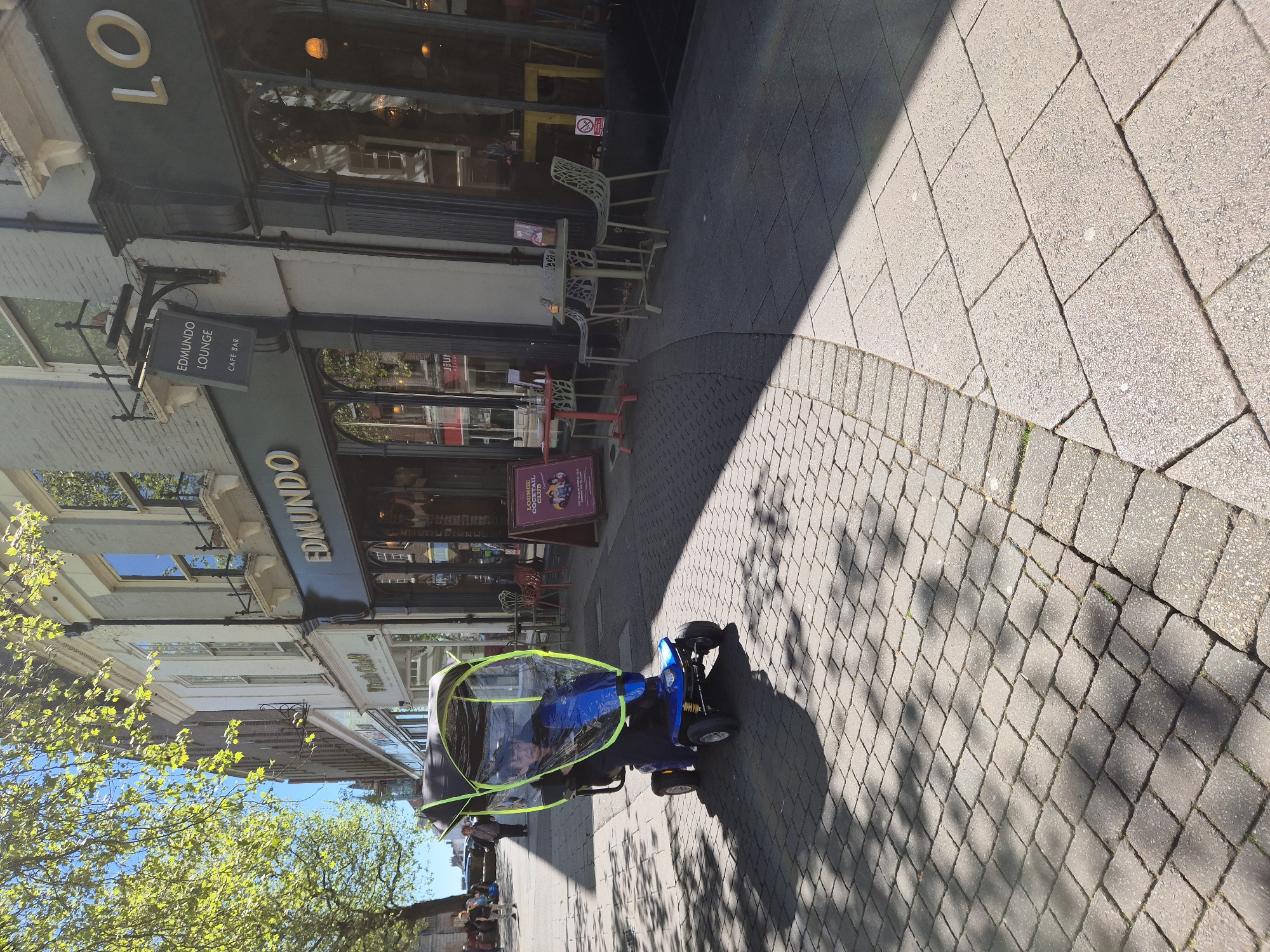 The image depicts a mobility scooter on a brick-paved sidewalk in front of a building. The building is identified by a sign that reads "EDMUNDO LOUNGE CAFE BAR." The location is in West Suffolk, United Kingdom. The scooter has a blue frame and a transparent rain cover trimmed with green. Seated on the scooter is a person wearing dark clothing. Further down the sidewalk, a few people are visible. The sky is partly cloudy, and there are trees with bright green foliage on the left side of the frame. The sidewalk is partially shaded. The overall scene suggests a typical day in a West Suffolk commercial area.
