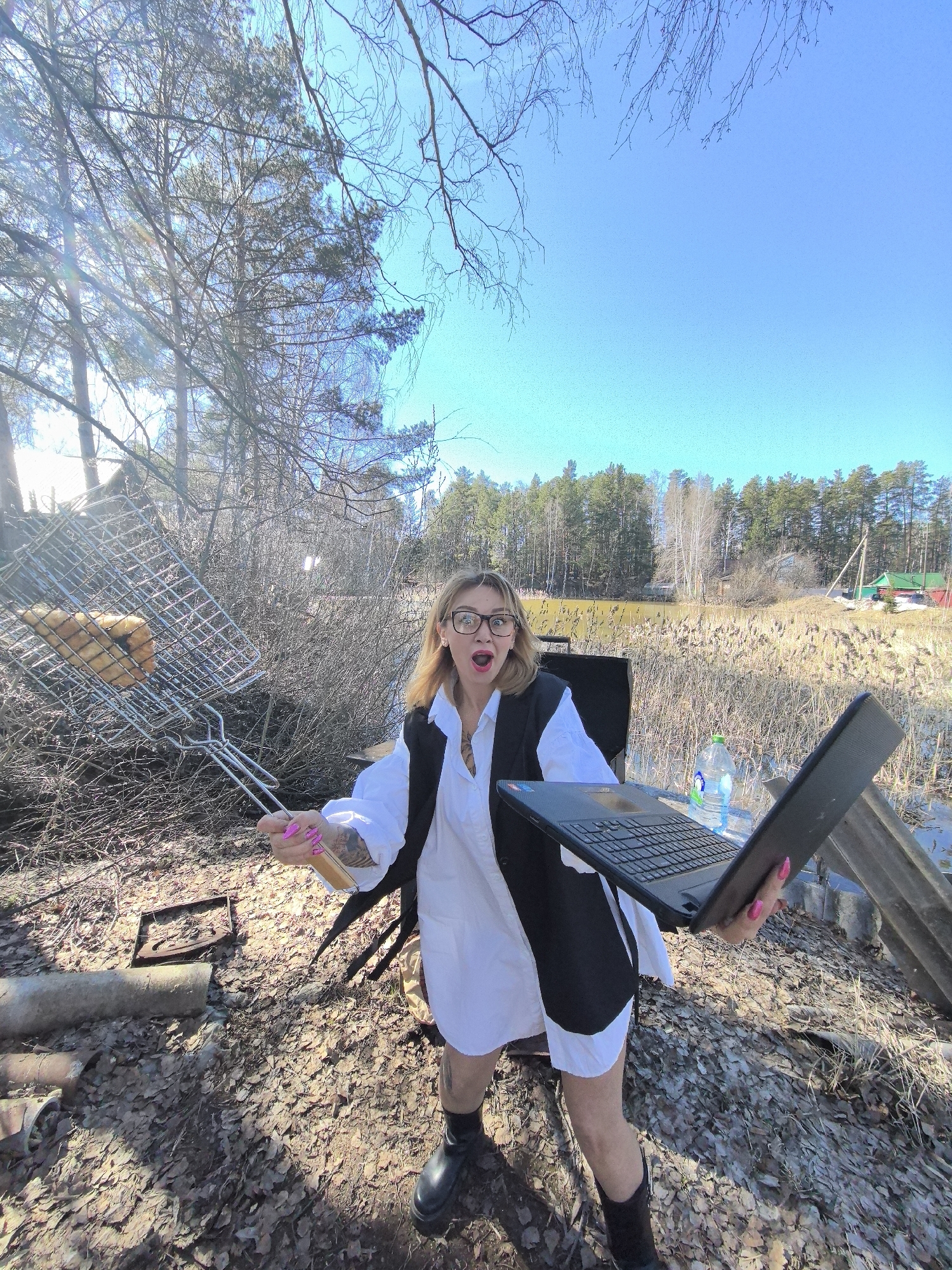 A woman stands outdoors on dry leaves and brush, holding a laptop in her right hand and a grill filled with what appears to be meat in her left. She is dressed in a white shirt and a black vest, wearing black boots and glasses, and she has tattoos visible on her arms and legs. Her mouth is open in an expression of surprise or excitement. Behind her is a wooded area with a lake or marsh, and in the background, one can see the edge of a settlement. A water bottle rests on a table behind the woman, and there is a chair also visible. The location of the image is in Noviy, Russia.