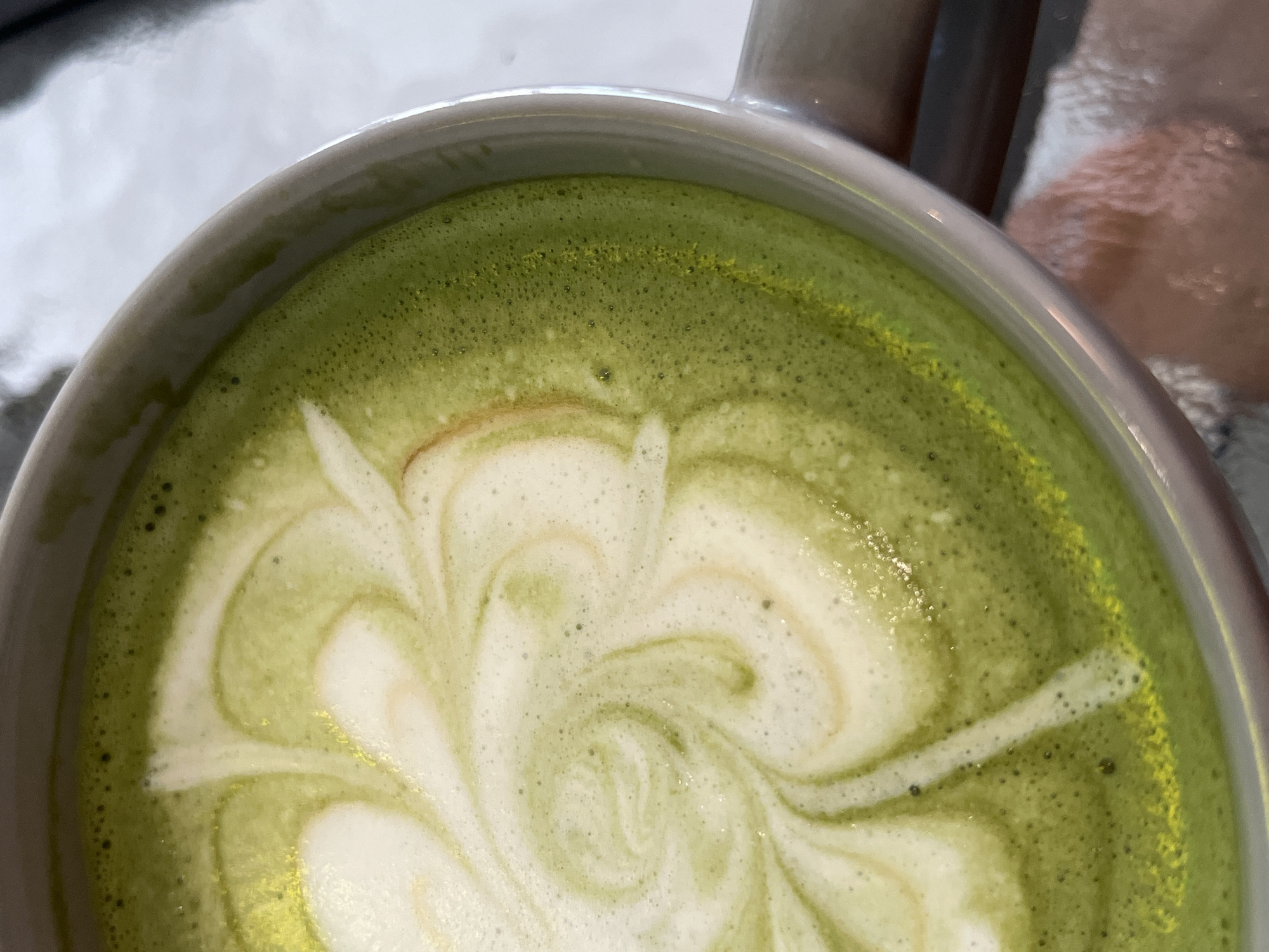 A close-up, eye-level shot reveals a cup of matcha latte with latte art. The latte is predominantly green, with white swirls creating a decorative design. The cup appears to be ceramic, with a gray or off-white color. The surface underneath is reflective. The location is Unknown, Indonesia.