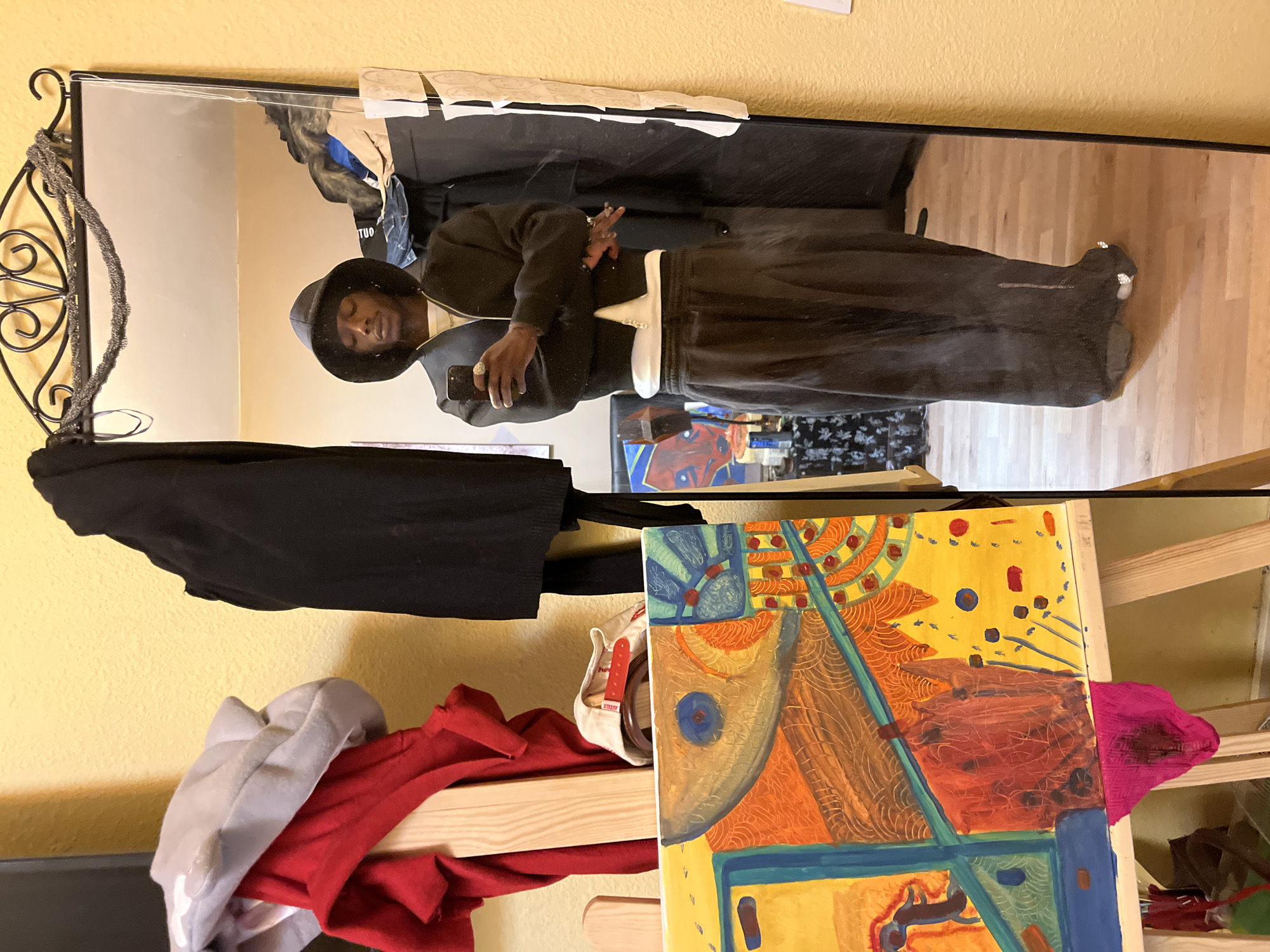 A person in black clothing and a hat is reflected in a mirror. The person is holding a smartphone, seemingly taking a picture of themselves. A painting on an easel is partially visible in the foreground. There are also clothes hanging on a rack or stacked nearby. The scene appears to be indoors, possibly in a bedroom or studio. There is no information suggesting the location is in Barcelona, Spain.
