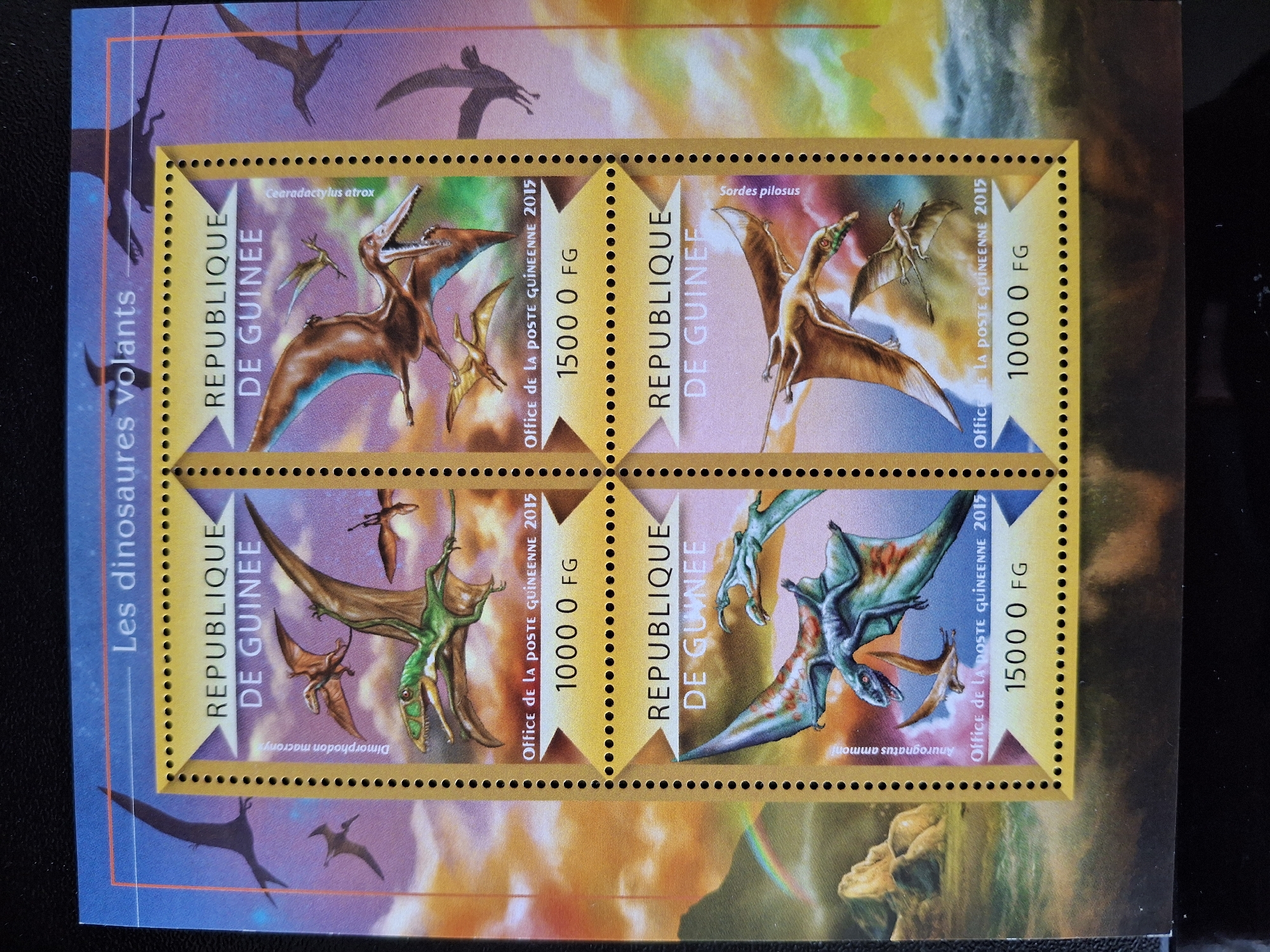 The image is of a sheet of postage stamps from the Republic of Guinea, issued in 2015, featuring illustrations of flying dinosaurs (pterosaurs). The sheet contains four stamps arranged in a 2x2 grid. Each stamp depicts a different species of pterosaur: Cearadactylus atrox, Sordes pilosus, Dimorphodon macronyx, and Anurognatus ammoni. The stamps also feature the text "REPUBLIQUE DE GUINEE", "Office de la POSTE GUINEENNE 2015", and the face value in francs (1000 or 1500 FG). The stamps are bordered by a yellow, dotted pattern. The sheet is titled "Les dinosaures volants" and is partially placed on a background depicting additional flying dinosaurs in silhouette and a landscape with a rainbow. There is no information about Pleven, Bulgaria.