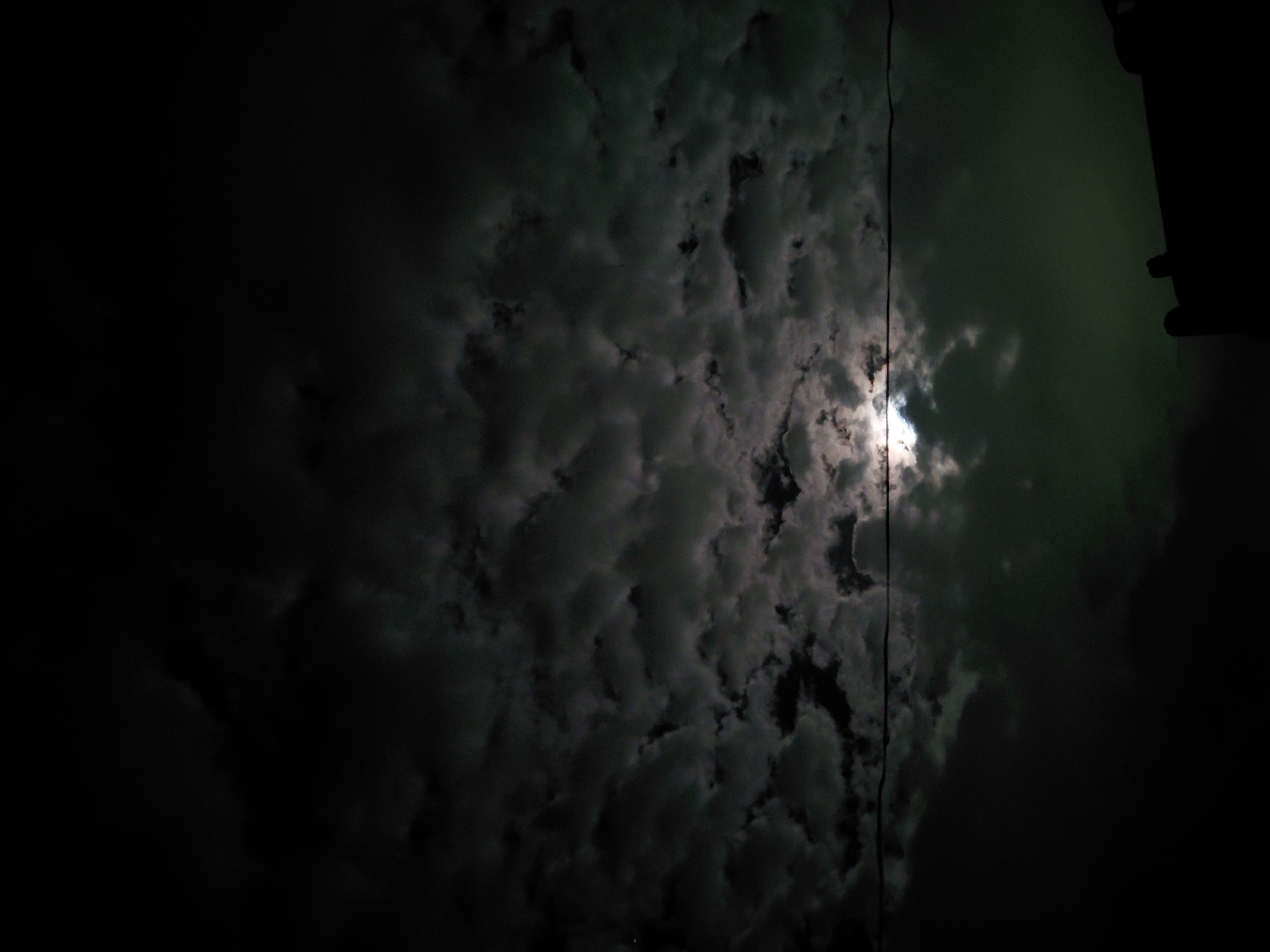 The image captures a dark, cloudy night sky, taken with an upward perspective. The primary subject is an expansive canopy of dense, dark grey clouds, which dominate the frame and exhibit varied textures and depths. A bright, circular light source, consistent with the moon, is visible through a break in the clouds on the right side of the image, casting a soft, luminous glow that illuminates parts of the cloud formations around it.
A thin, dark vertical line, appearing to be a cable or wire, runs downwards from the top edge, positioned just to the right of the center. On the far right edge, a dark, indistinct silhouette of what appears to be a building or structure is partially visible, suggesting an urban or semi-urban setting in Santa Ana, Venezuela.
The time of day is clearly night, with the moon being the main source of light breaking through the cloud cover. The weather is overcast. The color palette is composed mainly of deep blacks, dark greys, and some lighter greys where the moonlight penetrates, with a subtle greenish tint in the darker sky areas away from the moon. There is no discernible activity taking place, and no visible text can be identified in the image.