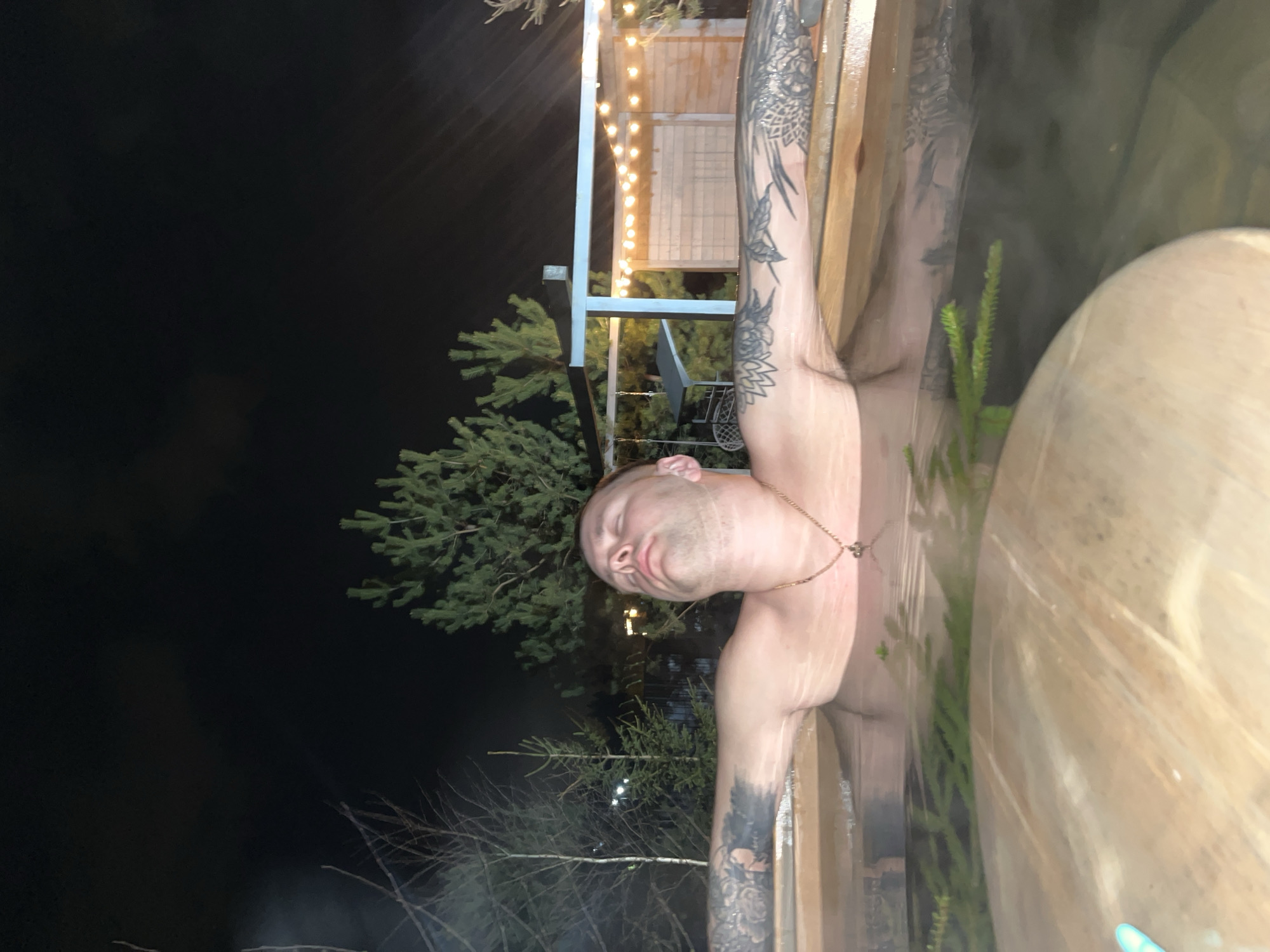 The image shows a shirtless man, likely in a hot tub or barrel sauna, outdoors at night. His head is tilted back, eyes closed, and he wears a necklace. His arms rest on the wooden edge of the tub, revealing tattoos on both arms. Evergreen branches and trees are visible in the background, along with lights strung on a structure. The location is possibly in Литвиново, Russia, based on the prompt.