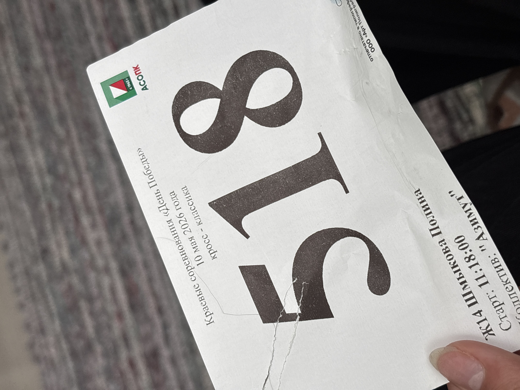 A close-up shot shows a printed ticket, bearing the number 518 in large font, in a hand. The ticket denotes participation in the "Day of Victory" regional competition, a cross-country classic race, to be held on May 10, 2026. The "ASOPK" logo and the company’s founding year of 1963 are printed on the top left. The ticket also provides the participant's name, Shmykova Polina, categorized as Ж14, with a start time of 11:18:00, representing the "Azimut" team. The ticket indicates it was printed at "Art Polygraph" LLC. The photograph appears to be taken in Perm, Russia.