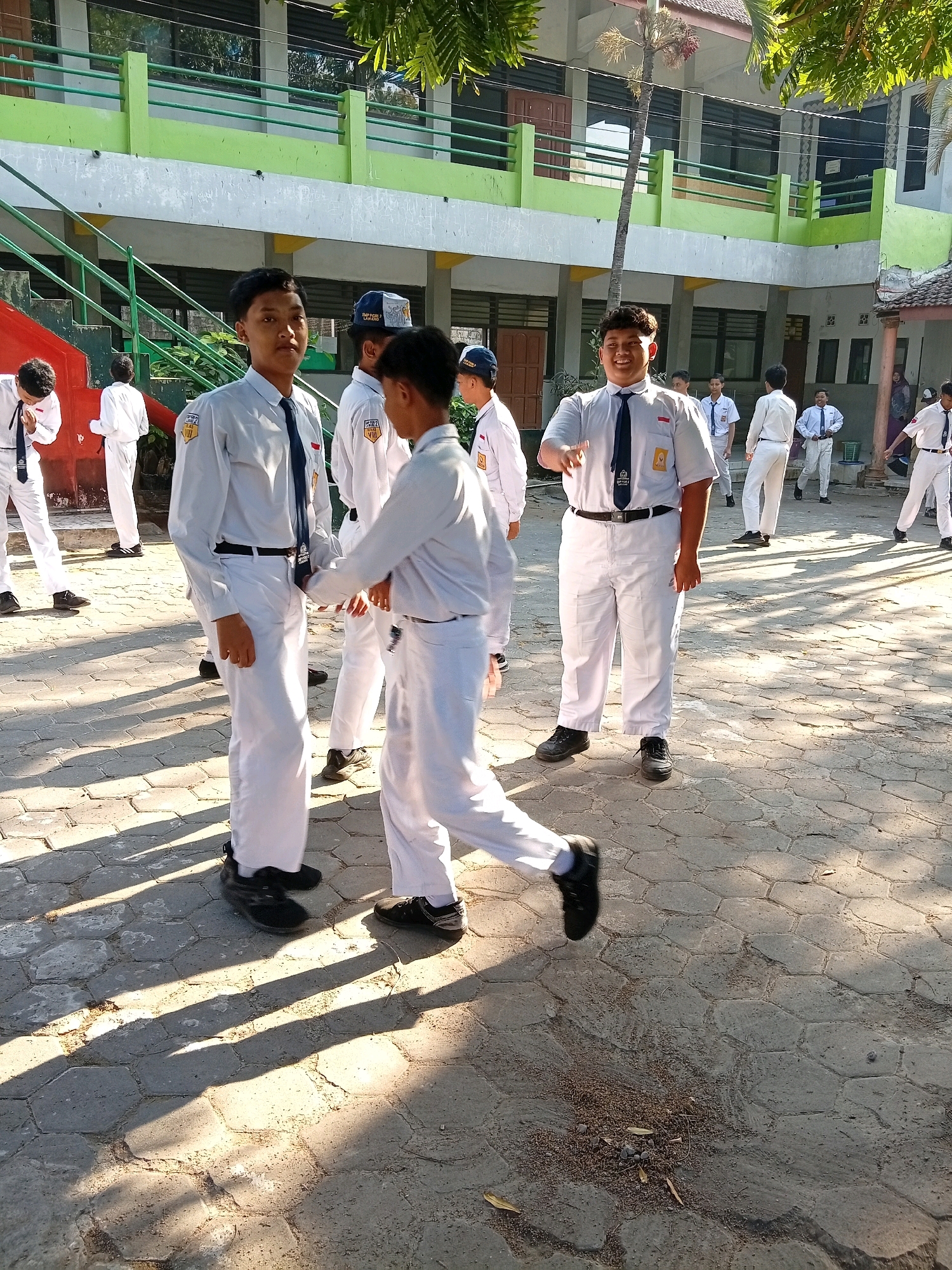 The image captures a group of male students in white school uniforms and ties standing in an outdoor courtyard. The architecture suggests a school setting, with a building featuring green and gray painted facades, balconies, and staircases visible in the background. The ground is paved with hexagonal stones, and sunlight casts distinct shadows, indicating a sunny day. The students appear to be in casual conversation or interaction, with some looking at the camera. The presence of palm trees and the architectural style suggest a tropical or subtropical location, consistent with Lawang, Indonesia. The overall atmosphere is that of a school break or a social gathering among students.
