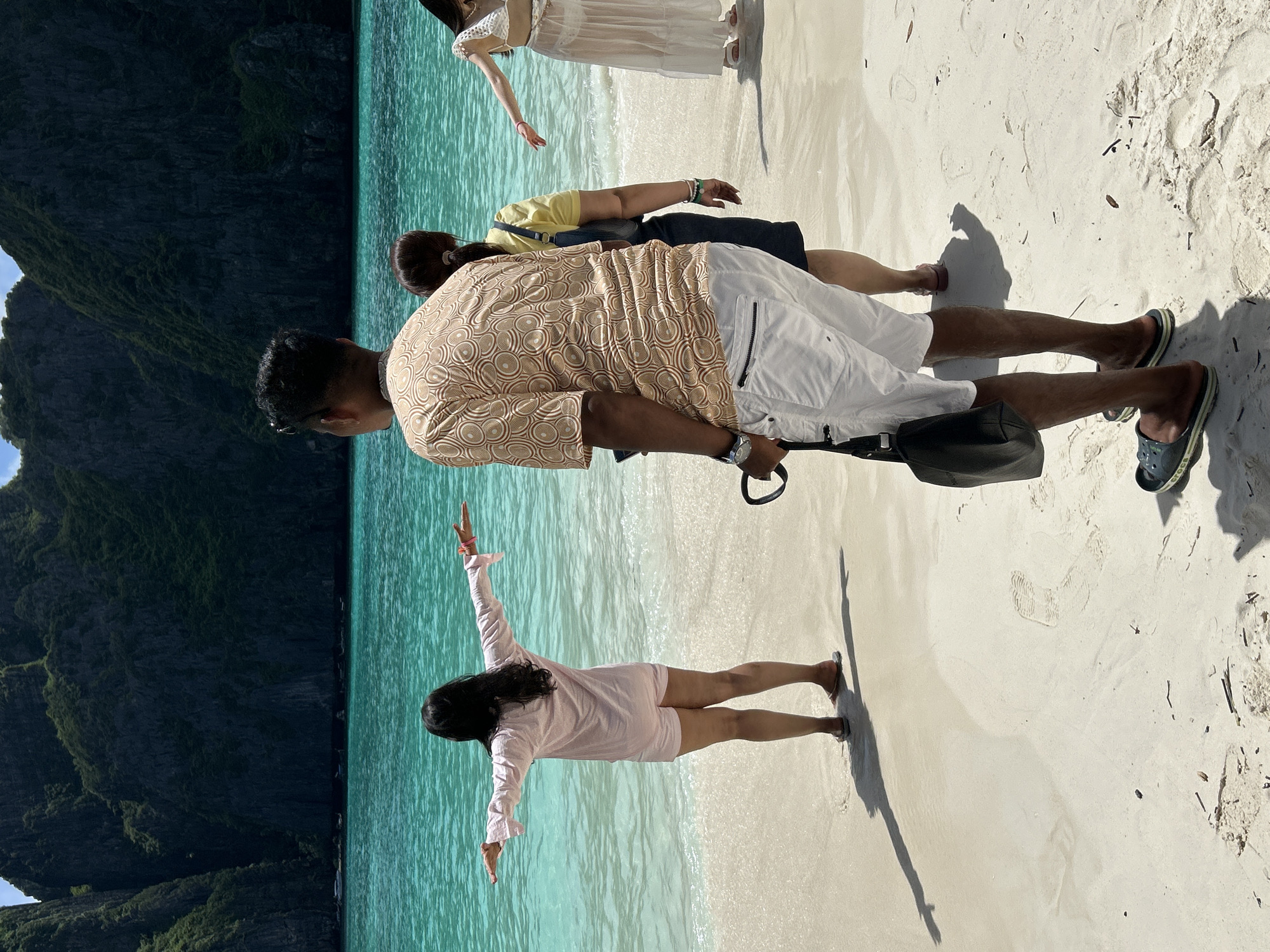 In Ao Nang, Thailand, on a sunlit beach with turquoise waters and towering limestone cliffs in the background, four individuals are present. A woman in the foreground, from a rear perspective, stands with her arms outstretched to her sides, wearing a light pink top and shorts. Beside her, a man faces away from the camera, dressed in a patterned brown and beige shirt and white shorts, carrying a black bag. Behind him, another woman in a yellow top and dark skirt walks away, and a woman in a white bikini and a sheer white cover-up stands further down the beach. The sand is a pale beige, with footprints and scattered debris. The water is clear and calm, with small waves lapping at the shore.