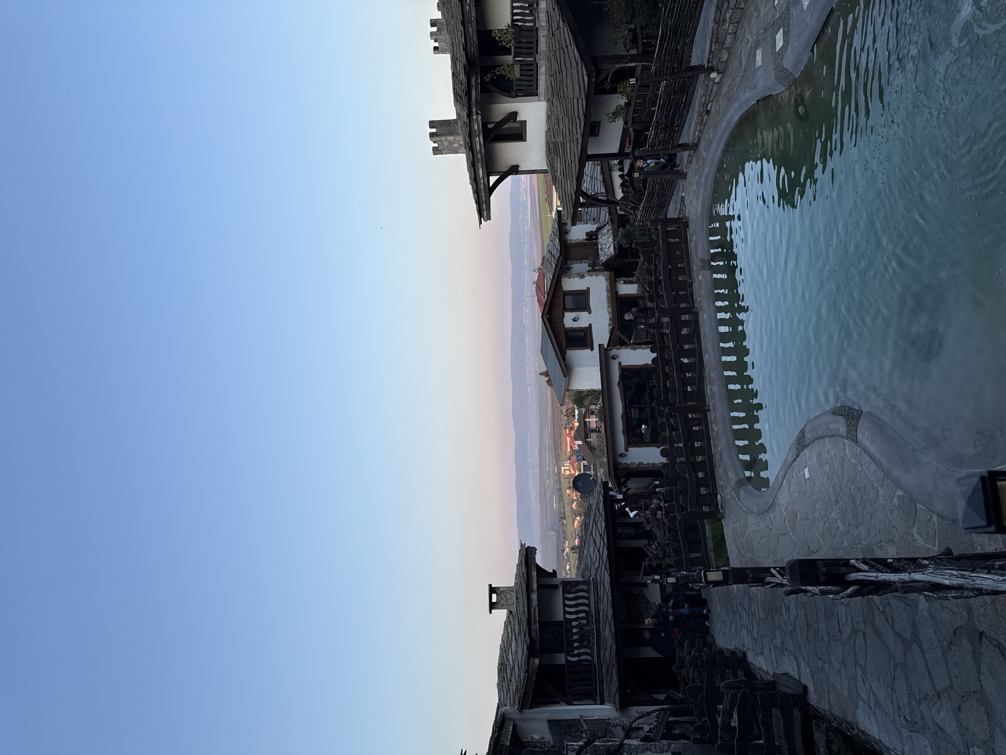 A vertically-oriented, angled outdoor shot depicts a building complex in Medovo, Bulgaria, with a stone-edged pool of water at the right. The scene is illuminated by the soft, ambient light of dusk or dawn, casting a subtle gradient of color across the sky.
The architecture consists of multi-storied, possibly traditional buildings with dark, stone-like shingle roofs and white walls. Dark wooden balconies and fences line the facade facing towards the pool. The pool itself is filled with opaque light-blue water, reflecting the ambient light.
In the background, a distant landscape with light green and brown hues suggests a valley or plain, bordered by faint mountains and a horizon where the sky meets the land. No people are clearly discernible within the frame, but a gathering of indistinct forms in dark clothes can be seen under an overhang. The environment is a blend of constructed elements and natural scenery.