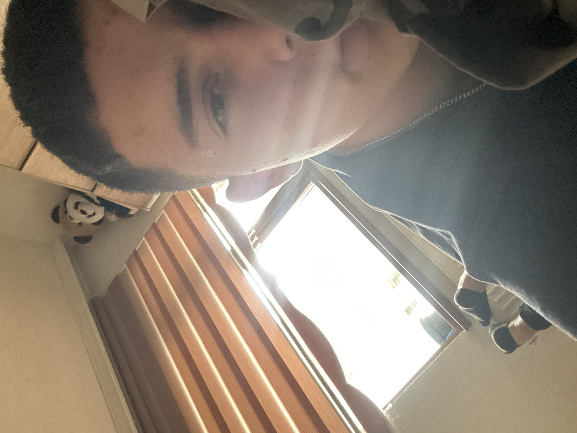 A selfie, taken at a low angle, features a person with short, dark hair and light skin looking directly at the camera. The person is wearing a dark shirt and a silver chain is visible around their neck.
Behind the person, a window with brown wooden blinds allows bright sunlight to enter the room. Outside the window, buildings can be seen in the distance, potentially indicating an urban area. The location is Zlatograd, Bulgaria.
To the left of the window, a panda plush toy is visible in the corner of the room, suggesting the presence of personal belongings. The lower part of the frame also shows the person's feet, in shoes and socks, up against the window frame.