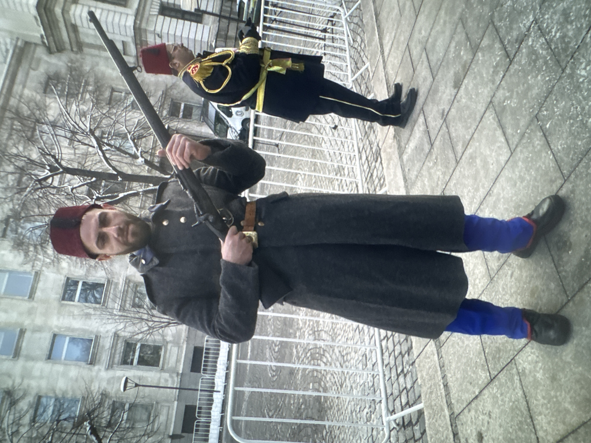This media file captures two men in ceremonial uniforms standing outdoors in what appears to be an urban setting. The image is oriented vertically, showing the subjects at an angle.
The man in the foreground, positioned on the left side of the frame, wears a dark grey long coat over bright blue trousers, black shoes with red detailing near the ankles, and a red fez hat. He has a slight beard and is looking directly towards the viewer with a neutral expression. He holds an old-fashioned rifle with a wooden stock, secured by a brown leather strap across his body. A golden buckle is visible on his belt. Bare tree branches are behind him, suggesting winter or early spring.
The man in the background, positioned further right, wears a dark blue uniform adorned with elaborate gold braiding on the chest and shoulders, a yellow sash or belt, dark trousers with a prominent yellow stripe, and black shoes. He also wears a red fez hat and is holding a rifle, though it is less distinct. He stands in a formal, possibly at-attention pose.
Both men are separated from the viewer by a white metal barrier, commonly used for crowd control or defining a parade route. They stand on a grey paved surface, consisting of large square tiles. In the background, there is a multi-story building with numerous windows, suggesting a city street or square. A street light pole is also visible.
The scene depicts a daytime event, likely a parade, ceremonial guard duty, or historical re-enactment, given the distinctive uniforms and rifles. There is no visible text in the image. The general atmosphere is formal and historical.