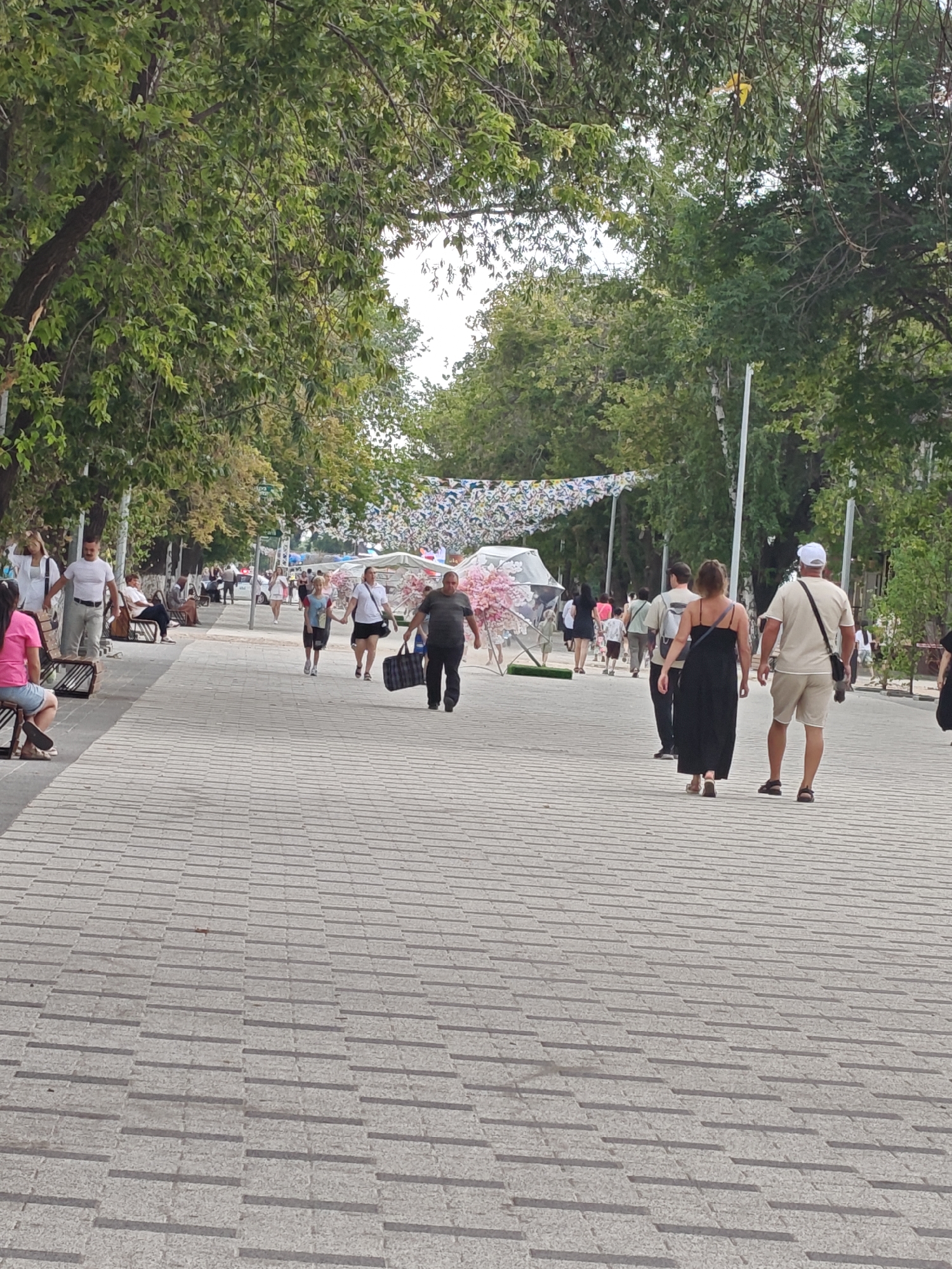 This is a full-body, eye-level, candid shot of a paved pedestrian walkway in the city of Petropavl, Kazakhstan. The walkway is lined with trees and appears to be in a park or public square. In the foreground, the walkway is paved with light-colored, rectangular stones arranged in a pattern. Several people are walking on the path. On the right side of the walkway, a man in a beige t-shirt and shorts is walking away from the camera, wearing a white baseball cap. A woman in a long black dress is walking in front of him. On the left side of the walkway, a woman in a pink shirt and shorts is sitting on a bench. Further down the walkway, a group of people are walking, including a man in a dark shirt carrying a large bag. In the background, there are more trees and some decorative elements hanging over the walkway, possibly part of a festival or event. The lighting suggests it is daytime, possibly late afternoon due to the shadows.