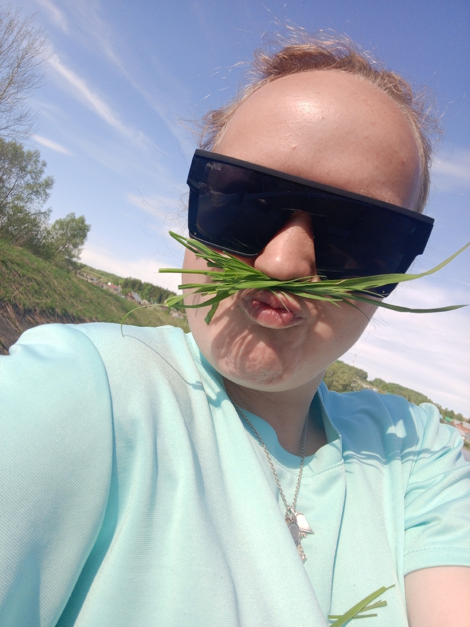 A person with light skin, wearing dark sunglasses and a light blue shirt, is holding green blades of grass under their nose, resembling a mustache. They are outdoors, with a blue sky and light clouds in the background. Green foliage and a building or structure are visible in the distance. The person is also wearing a necklace with a pendant. The location is Уральское, Russia.