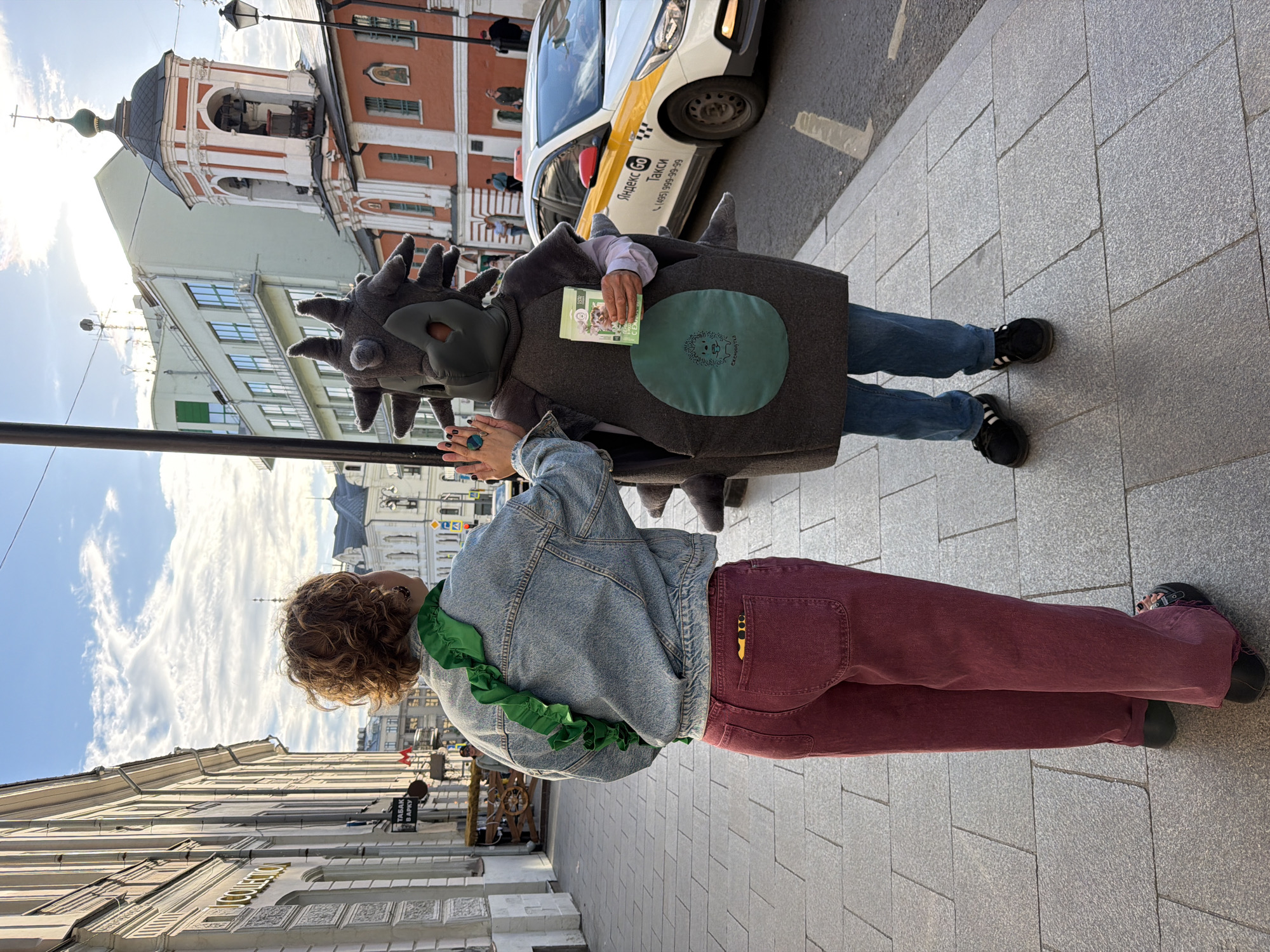 A person dressed in a gray hedgehog costume stands on a city sidewalk next to a person in a denim jacket and maroon pants. The "hedgehog" holds a package of cat food. A white Yandex Taxi car is partially visible in the background. Architecture in the background indicates a location in Moscow, Russia. The church of St. Nicholas is visible behind the "hedgehog."