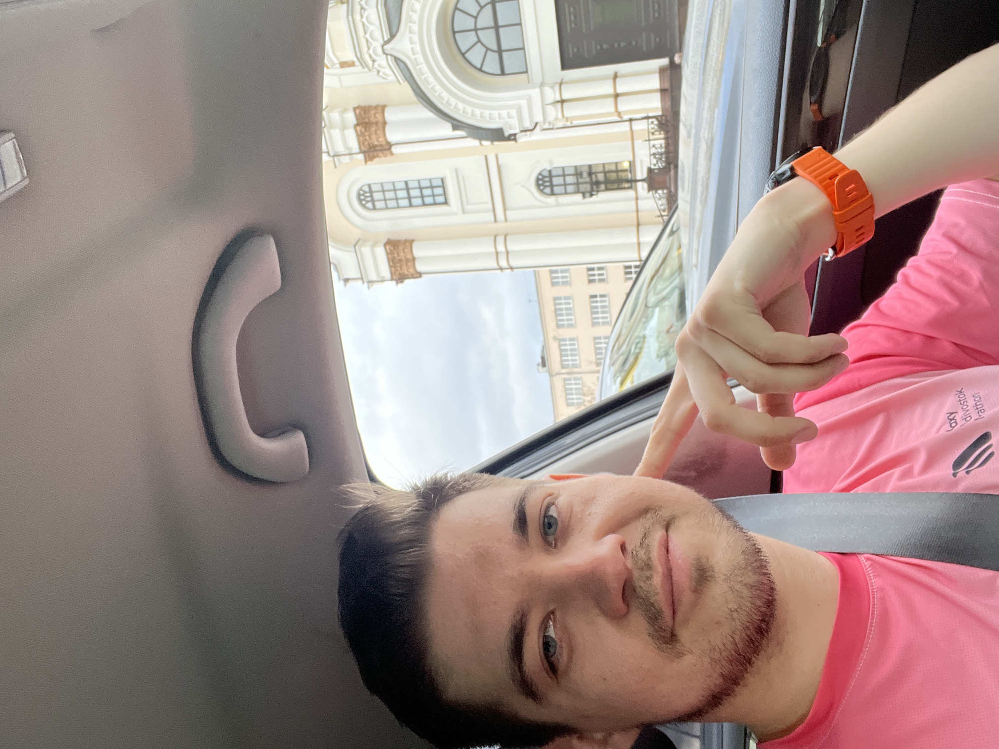 A man wearing a pink shirt and orange watch is taking a selfie inside a car. He has short dark hair, a light beard, and blue eyes. He's wearing a seatbelt. Outside the car window, there is a building with a white facade and arched windows. The building is in Yekaterinburg, Russia.