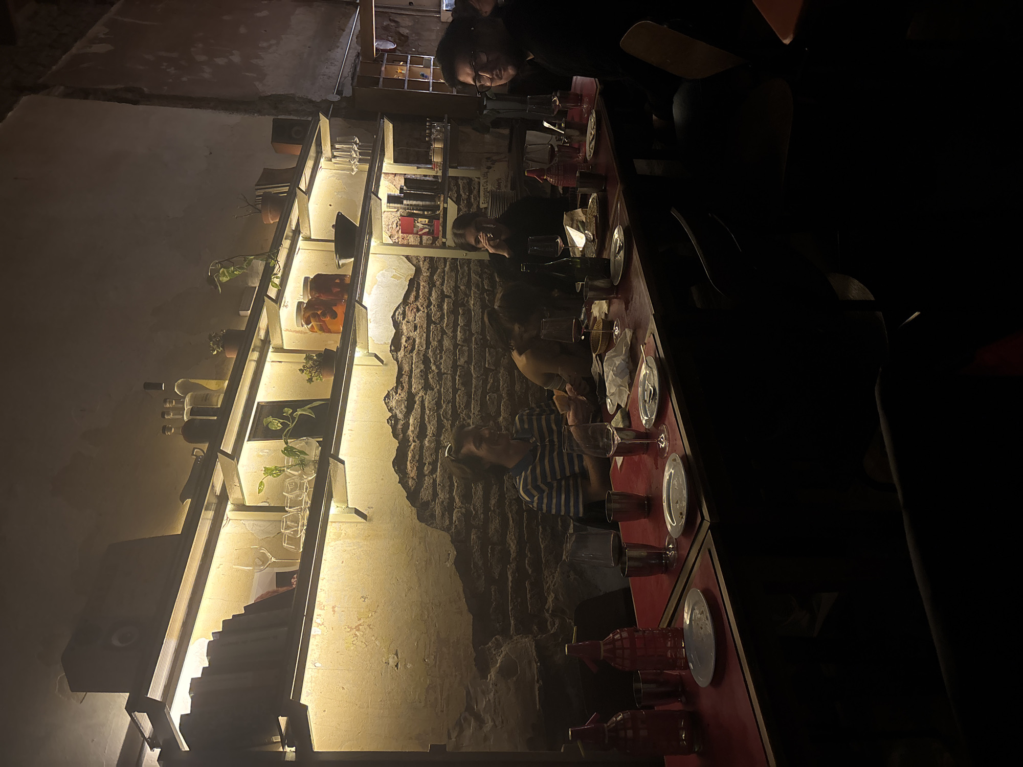 The image captures an interior scene, likely a restaurant or bar in Buenos Aires, Argentina, at night or in the evening, characterized by dim, warm lighting. The perspective is angled downwards, looking along a long, narrow dining table.
Several people are seated along one side of the reddish-brown table. Their faces are partially obscured or dimly lit. One individual, wearing glasses and a dark top, looks towards the viewer. Further down, another person in a blue and white striped shirt is visible, engaged with their companions. The people appear to be dining or socializing, with plates, various glasses (including wine glasses and tumblers), and small condiment bottles on the table. In the foreground, red, patterned bottles, possibly seltzer bottles, are prominent.
Along the wall parallel to the table, two long, illuminated shelves display an array of objects. These include small potted plants, clear and darker bottles (suggesting beverages), stacks of books, and more glassware. The shelves are lit from below, casting a warm glow onto the textured wall behind them, which appears to be made of rough stone or brick in some sections, and a smoother, lighter material in others. The overall ambiance is intimate and cozy, with the low light creating a rustic yet sophisticated feel. Behind the person in glasses, faint text on a box on the upper shelf reads "Express Co." and possibly "Milwaukee".