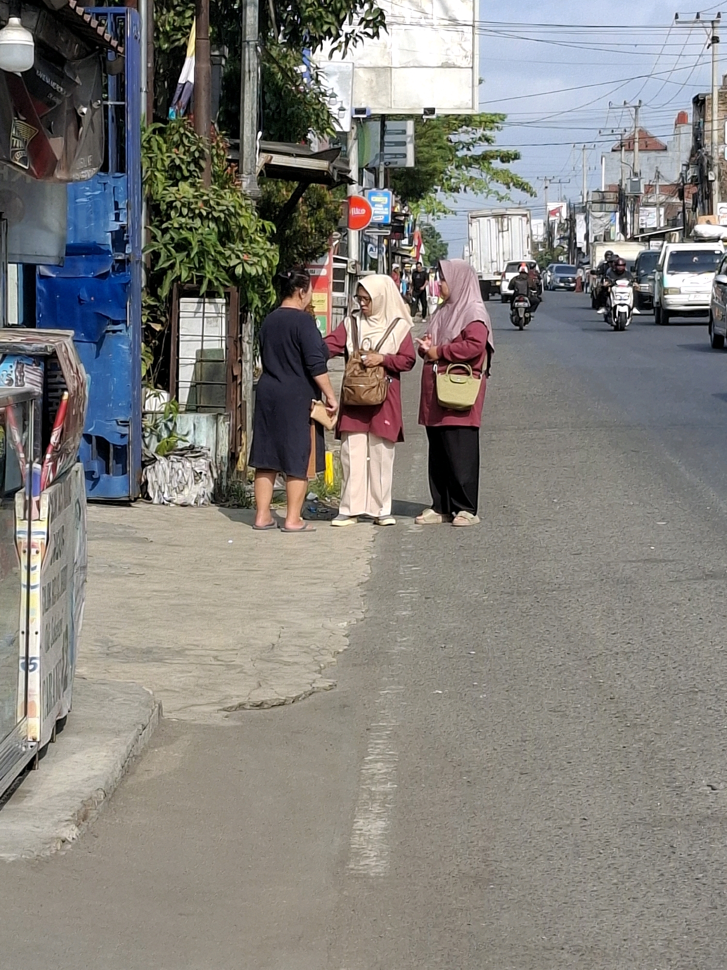 A medium-shot image captures a street scene in Sukaraja, Indonesia, with three individuals standing on a sidewalk. The person on the left, dressed in a dark navy dress and flip-flops, faces away from the camera and converses with two women. Both women are wearing hijabs and modest clothing. The woman in the center has a beige hijab and a pink-ish shirt with cream-colored pants, carrying a brown shoulder bag. The woman on the right wears a lighter pink hijab and a matching pink top with black pants, also carrying a small lime green bag. They are positioned close to each other, suggesting an ongoing interaction. The background reveals a busy street with traffic, including motorcycles and cars, indicating a daytime urban environment. Shops and buildings line the street, with some greenery visible. The sidewalk shows signs of wear and tear, with cracked pavement in some areas. The overall impression is of a casual street encounter in a developing urban area.