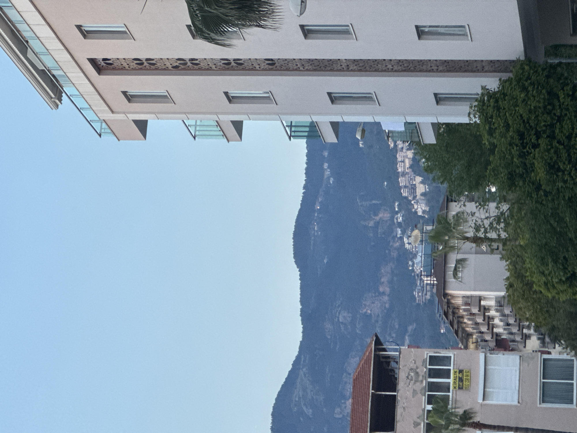 The image depicts a cityscape with a large mountain in the background. The foreground contains buildings with windows, including one with a "Kiralik" sign and a phone number. There are also palm trees visible. A taller building is visible at the top left, with a decorative band below its roofline. The sky is clear and blue. The exact location within an unknown city and unknown region cannot be determined from the visual information provided.