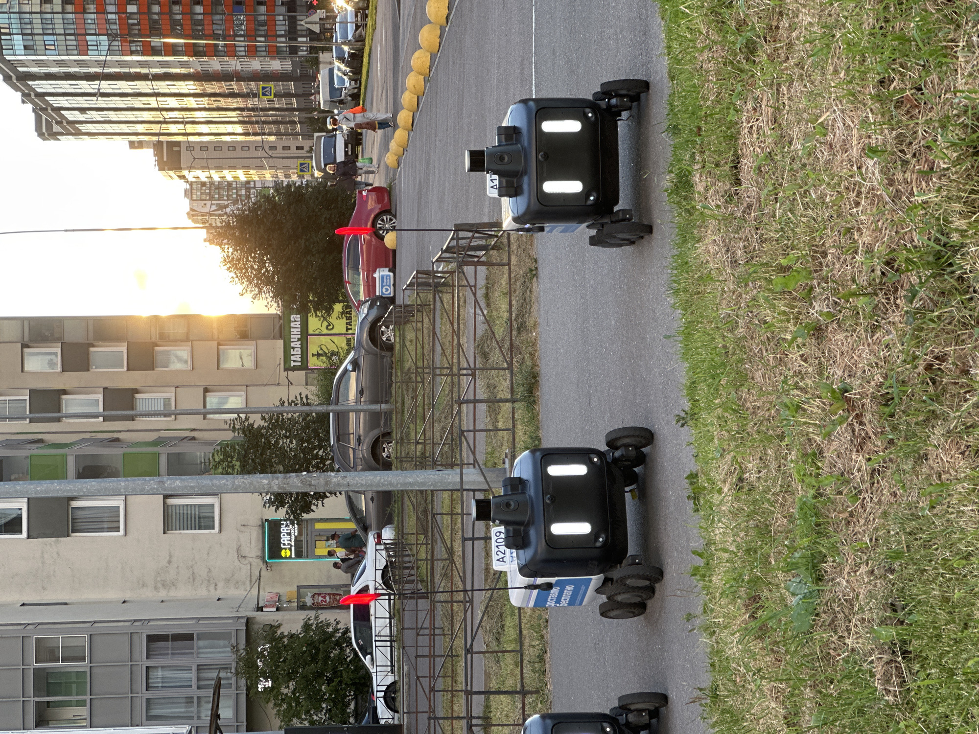 In Kudrovo, Russia, on a paved road next to a grassy area, three autonomous delivery robots are positioned. Two of the robots are in focus in the foreground, while the third is partially visible on the left. The robots are black and white, with illuminated front lights and wheels. One robot has a license plate that reads "A2109". A red flag extends from the top of one of the robots. In the background, several cars and buildings are visible. The buildings appear to be apartment complexes and commercial establishments. A street with parked cars and a sidewalk with yellow bollards is also present. The sun is setting, casting a warm glow on the scene.