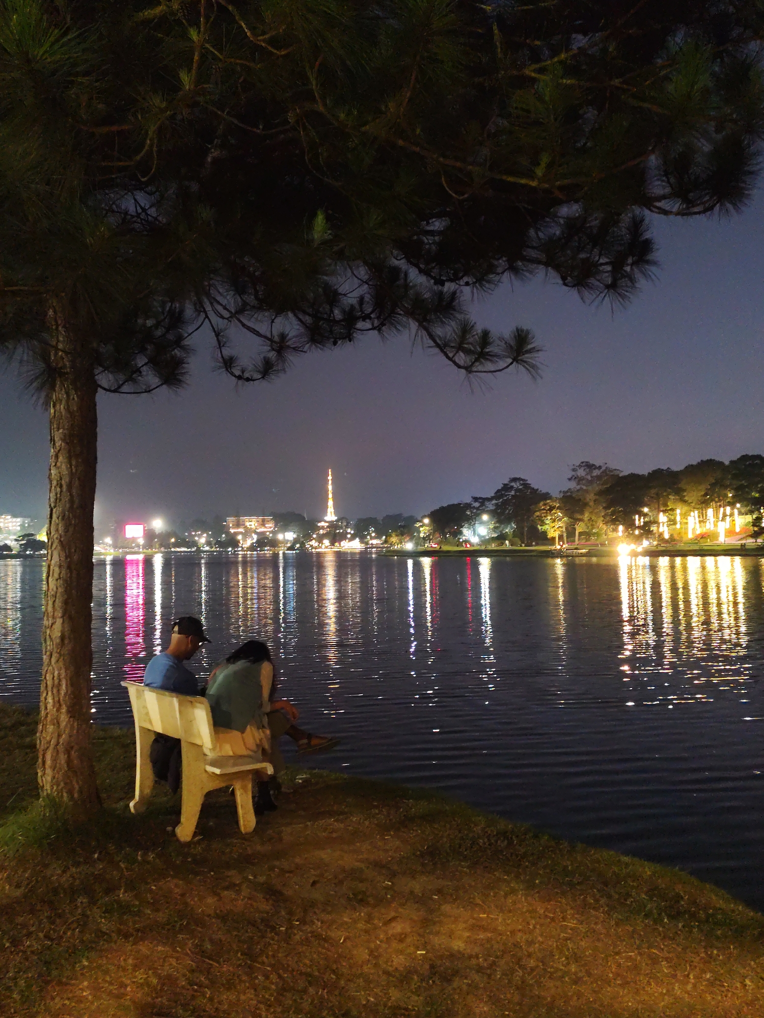 At night, a man and a woman sit close together on a white bench under a pine tree on the grassy shore of Xuan Huong Lake in Da Lat, Vietnam. Reflections of lights from buildings and the communications tower across the lake shimmer on the water.