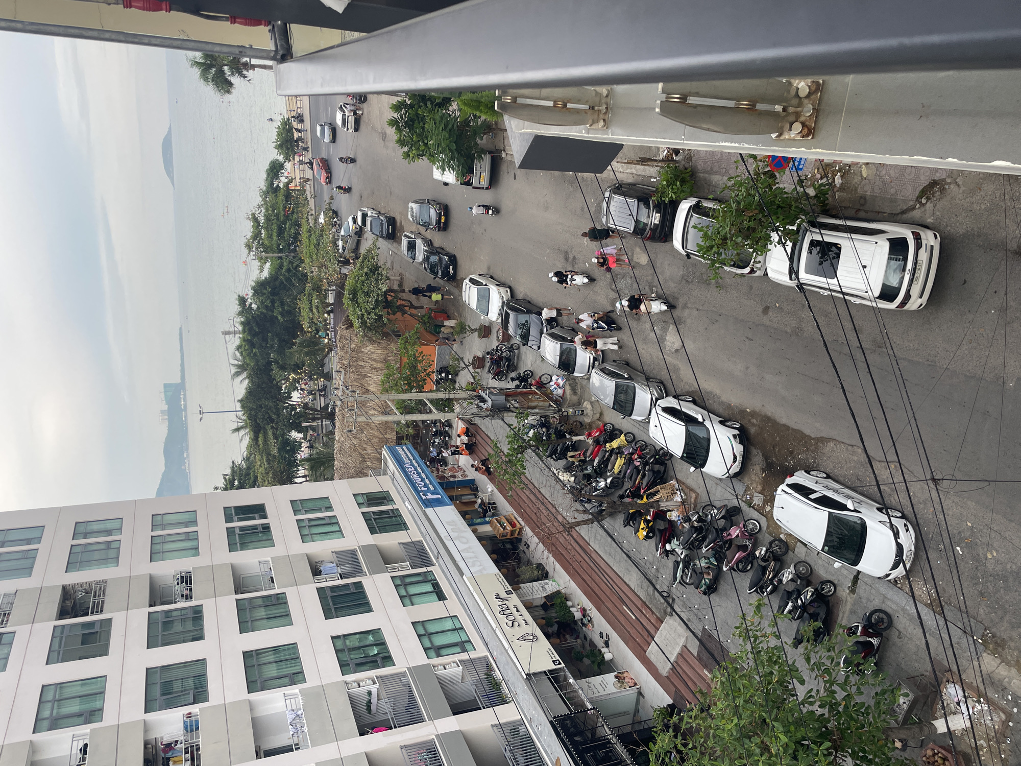 This is a high-angle, medium shot of a street in Nha Trang, Vietnam, near the beach. A tall, modern apartment building with many balconies and windows dominates the left side of the frame. Below the building, a row of businesses with awnings lines the street, including "Sofia Spa," "ASIA Spa," and "FOUR SEAS Restaurant." The street is filled with parked motorcycles and cars, and several people are walking on the sidewalks or riding motorcycles. In the background, palm trees, a beach, and the ocean are visible, with a few boats on the water and islands in the distance. The sky is overcast.