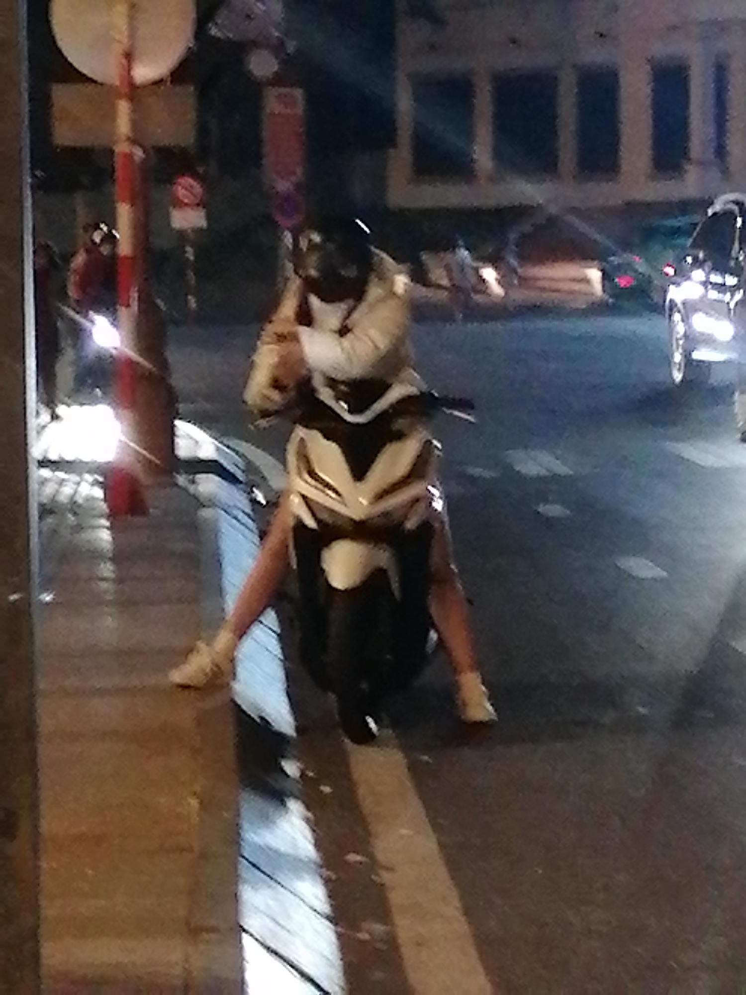 An image taken at night on a street in Da Lat, Vietnam shows a person on a scooter. The person is wearing a helmet and a light-colored jacket. Their legs are straddling the scooter, and their feet are on the ground. The scooter is black and white. In the background, there are buildings and other vehicles, including a car that appears to be a black sedan. The street is wet, suggesting it may have rained recently, and there are streetlights casting reflections. A red and white striped pole, possibly a traffic sign post, is visible on the left side of the image.