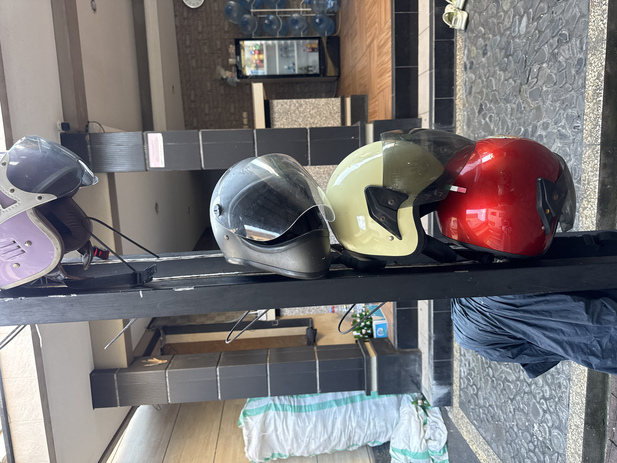 A vertical shot captures a rack with five motorcycle helmets hanging from a black metal pole, located outdoors in Kuta, Indonesia. The top helmet is light purple with a transparent visor. Below it is a black helmet with a transparent visor. The third helmet is light yellow with a dark tinted visor. The fourth helmet is also light yellow with a dark tinted visor. The bottom helmet is bright red with a black visor. A blue tarp is draped over the base of the rack. In the background, a white sack is visible to the left, and a building with a tiled floor, a glass-fronted refrigerator filled with drinks, and a rack of water bottles are to the right. The ground is paved with grey cobblestones and white gravel.