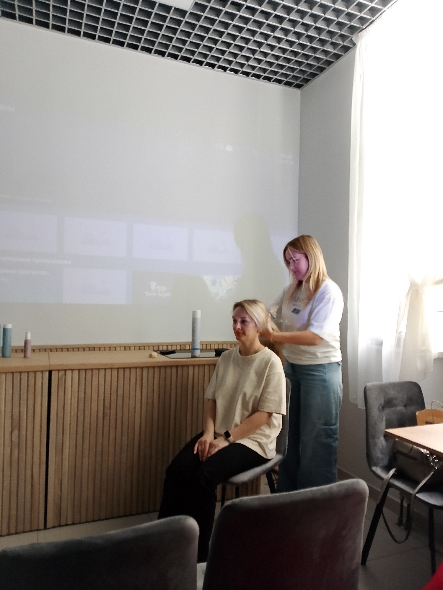 A woman with blonde hair is standing behind another woman, who is seated. The standing woman is arranging the hair of the seated woman. Both are wearing casual clothing. A wooden cabinet is behind the seated woman, with several items placed on top. A large screen is behind the cabinet, displaying visual content. The room has a drop ceiling with a grid pattern. Chairs are in the foreground and near a table. A window is visible in the background. This appears to be a room in Izhevsk, Russia, based on visual clues potentially present on the screen.