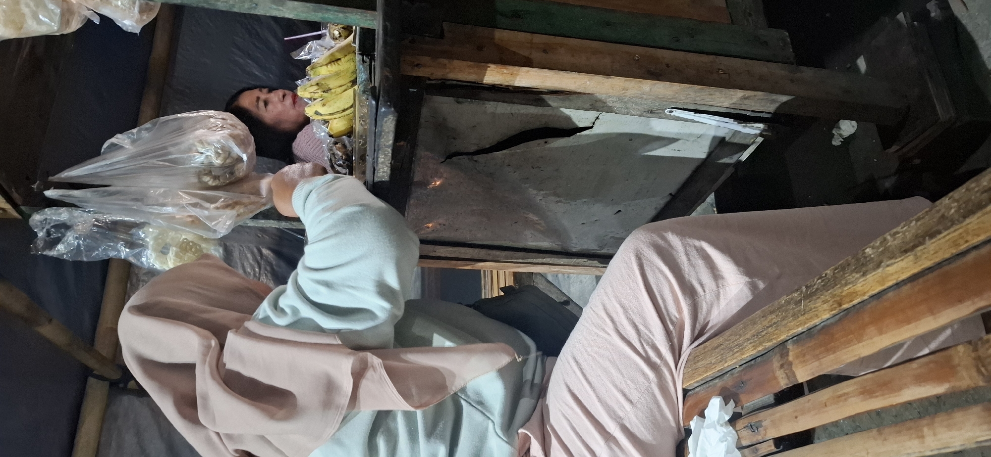 In Desa Gagaksipat, Indonesia, a person wearing a pale pink hijab and a light blue sweatshirt is seated on a bamboo bench, facing a makeshift stall. The stall is constructed with rough wooden planks and features a damaged, cracked surface on its front panel. A second person, a woman with dark hair, is visible behind the stall, looking to the left.
On the stall's surface, a bunch of ripe yellow bananas is arranged next to what appears to be a small serving of fried food. Several plastic bags filled with assorted items, likely snacks or food items, are suspended from the stall's overhead structure. The background is obscured by dark sheeting, and the lighting suggests an outdoor, possibly evening setting. A crumpled white tissue is on the bench to the left of the seated person.