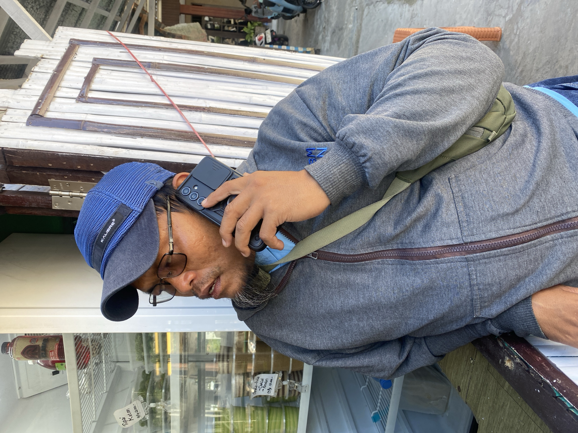 A man with a beard and glasses is standing and holding a cell phone to his ear, seemingly in conversation. He is wearing a blue baseball cap and a grey hooded sweatshirt. A green bag is slung across his chest. In the background, there is a white wooden door, a refrigerator filled with food containers, and a glimpse of motorbikes parked outside. The setting appears to be a street-side stall or shop. The image is taken outdoors in what appears to be a tropical climate, given the presence of a fan and bright sunlight.