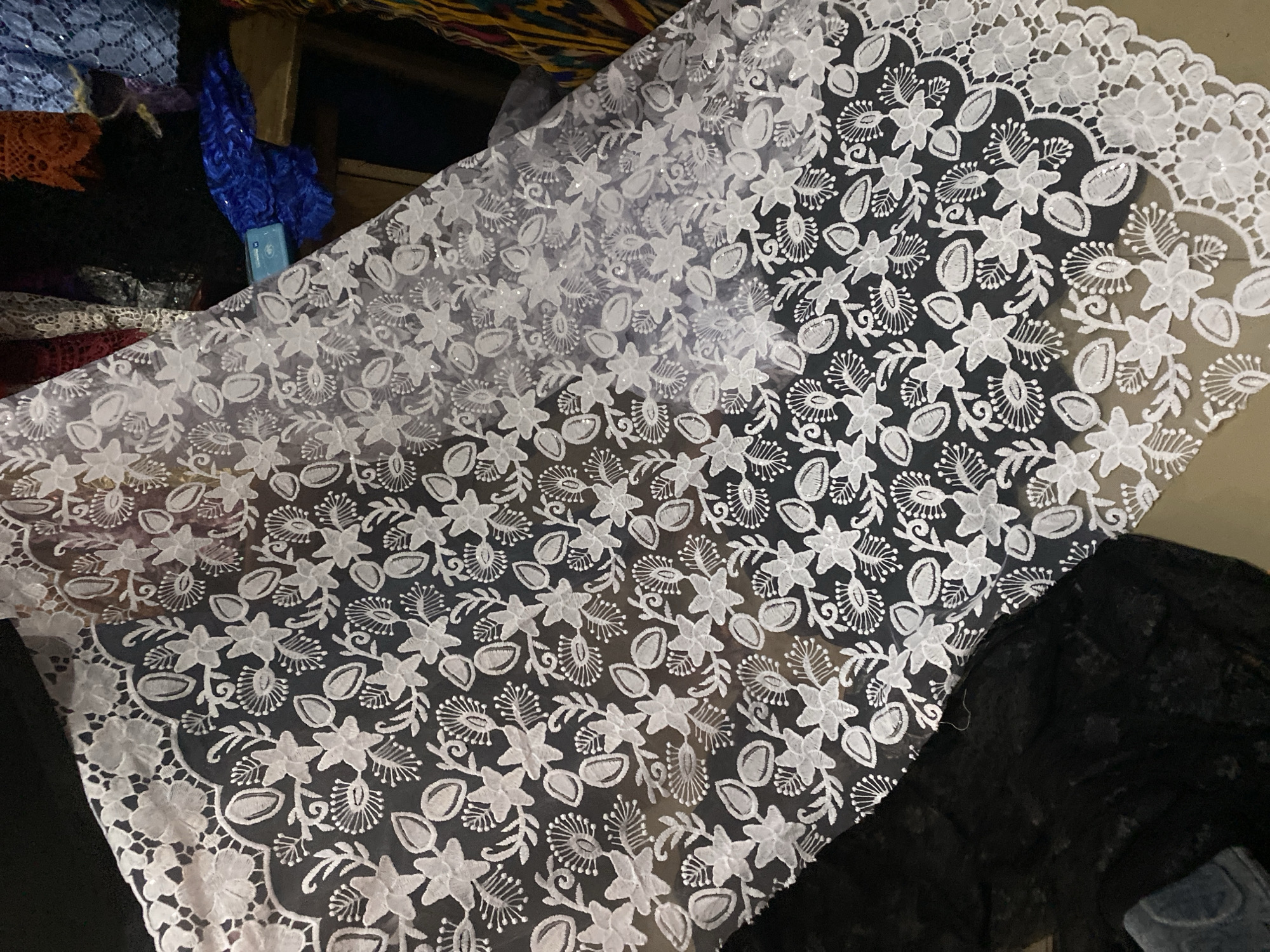 This media file features a close-up, high-angle shot of a white lace fabric with an intricate floral pattern. The fabric is draped and spread out, revealing its delicate texture and detailed embroidery. The pattern consists of stylized flowers with five petals, interspersed with smaller decorative elements resembling leaves and buds, all outlined in white. Scattered across the fabric are small, shimmering sequins that catch the light, adding a subtle sparkle. The lace appears to be laid over a dark surface, likely another piece of fabric, which provides a stark contrast and makes the white pattern stand out. In the background, hints of other fabrics are visible, including a vibrant blue fabric and possibly some patterned textiles, suggesting a market stall or a tailor's shop. The overall impression is one of fine craftsmanship and elegant design, commonly found in textiles used for clothing or decoration. There is no visible text in the image, and no specific time of day, weather conditions, or emotions are conveyed. The context of Jalingo, Nigeria, suggests this might be a locally made or imported textile available in the region.