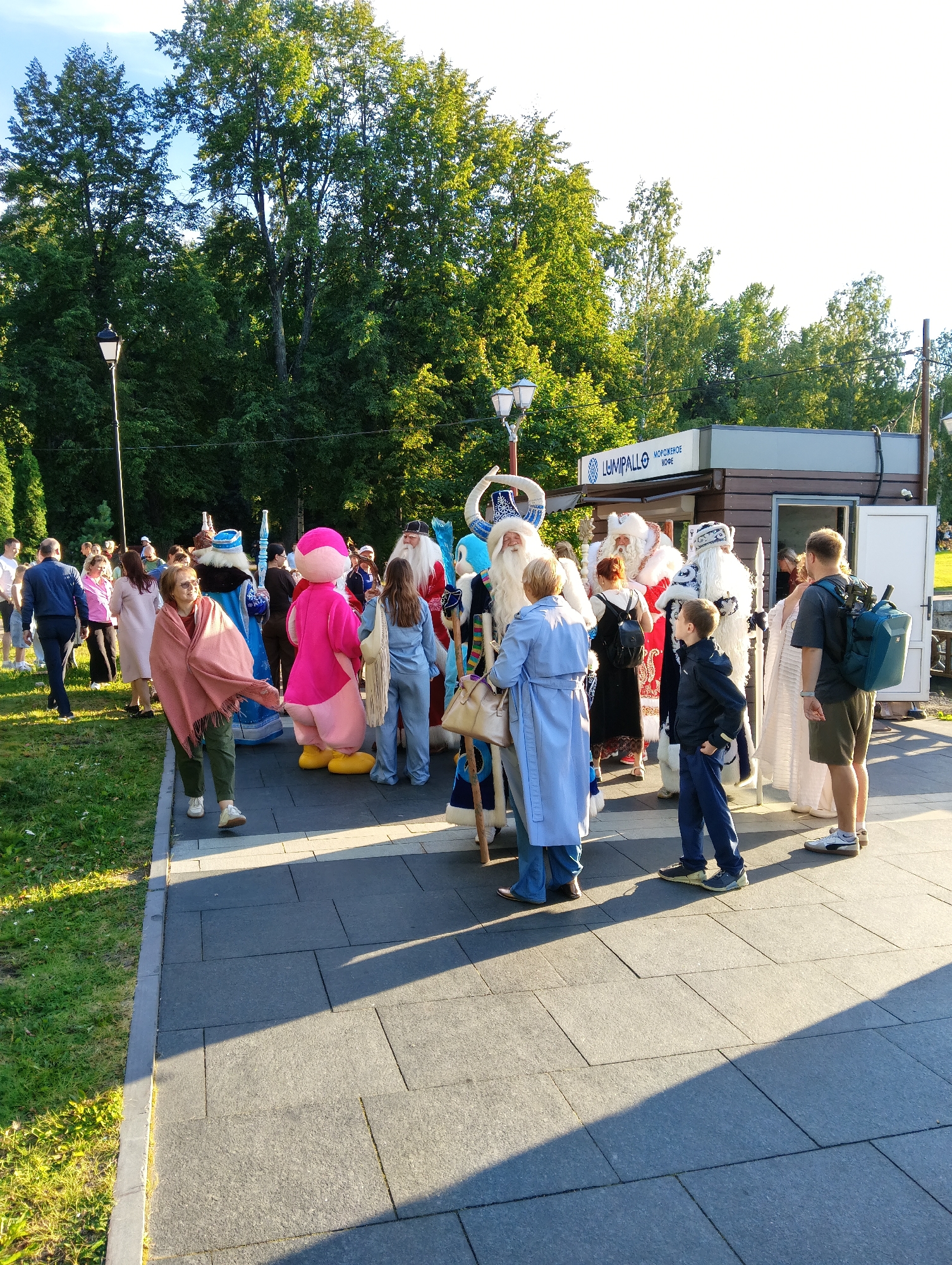 The image shows a sunny outdoor scene, likely in Petrozavodsk, Russia, featuring a gathering of people dressed in costumes, including multiple individuals dressed as Ded Moroz (Father Frost) and Snegurochka (Snow Maiden), traditional Russian winter characters. The costumes are elaborate, with one Ded Moroz in a red and gold outfit, another in a blue and white patterned robe, and a third in a white fur-trimmed costume. A person in a bright pink mascot costume, resembling a large bird or character, is also present. Several other individuals, some in everyday attire, are interacting with the costumed characters or observing the scene.
The setting appears to be a park or public square. In the background, there is a small kiosk with signage that reads "LUMIPALLO" and "МОРОЖЕНОЕ КОФЕ" (Ice Cream Coffee), suggesting a food or beverage vendor. A lamppost is visible to the left, and mature trees with lush green foliage line the background. The ground is paved with dark grey square tiles, and there is a strip of grass to the left. Sunlight casts long shadows across the paved area, indicating it is late afternoon. The overall atmosphere seems festive, possibly during a holiday event or celebration.