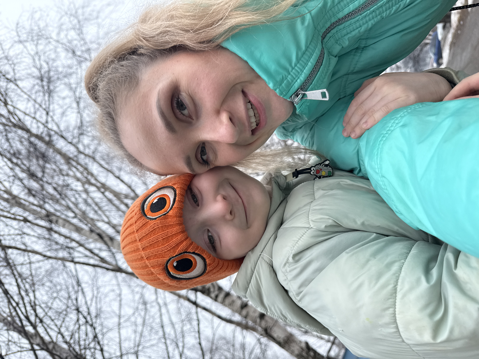 A photo taken in what appears to be an outdoor area of Chaikovsky, Russia, given the bare trees and overcast sky typical of the region. Two people are visible: an adult with blonde hair, wearing a turquoise winter jacket, and a child wearing an orange beanie with large eye designs and a light green winter jacket. They are posed closely, their faces near each other, smiling towards the camera. The background consists of bare tree branches against a grey sky.