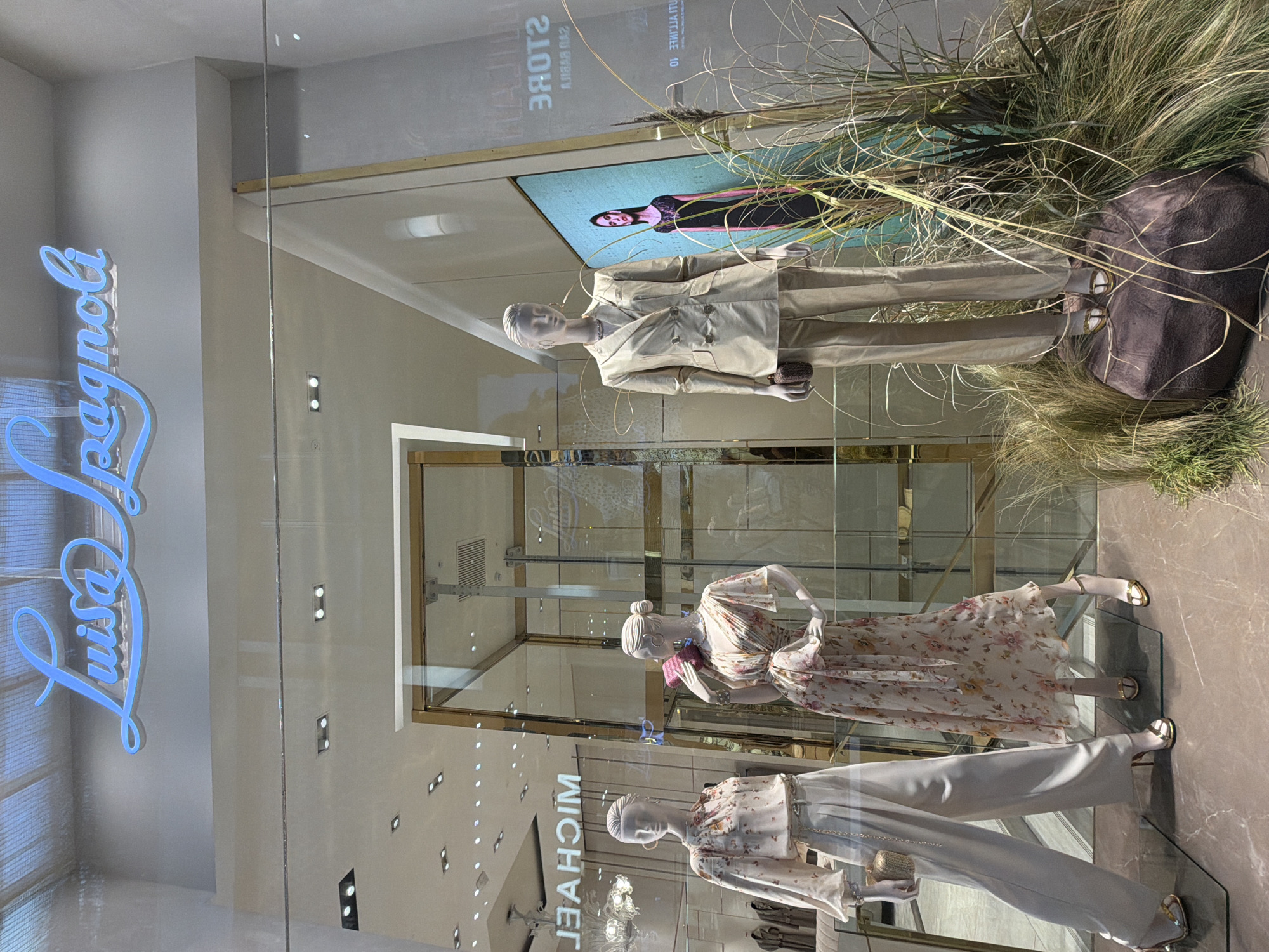 This image captures a brightly lit shop window display from a fashion boutique. Three female mannequins are showcased, dressed in contemporary attire, set against a backdrop of natural-looking dry grasses and an illuminated digital screen. The display is seen through glass, which reflects elements from the exterior and interior, including a prominent neon sign.
The uppermost mannequin wears a light beige or khaki-colored suit, comprising a blazer and trousers, and holds a small, dark, textured clutch bag. Behind it, a digital screen displays an inverted image of a woman in a black dress, with the reversed text "STORE" and "SPAGNOLI". To its right, a large, dark, organic-shaped prop is nestled among the tall grasses.
The middle mannequin is dressed in a flowing, light-colored dress adorned with a floral pattern of pink, yellow, and green hues, and has a small pink item draped over its arm. The lowest mannequin features light-colored, wide-leg trousers and a patterned blouse, accessorized with a small golden, textured handbag. All three mannequins have smooth, featureless white faces and wear elegant gold-colored sandals.
Reflected on the glass, a bright blue neon sign in cursive script clearly spells out "Luisa Spagnoli", indicating the brand of the boutique. Other reflections include internal lighting fixtures and the reversed word "MICHAEL" near the bottom, possibly from another store or internal signage. The overall impression is a stylish retail presentation, likely in Milan given the context, highlighting spring/summer fashion.