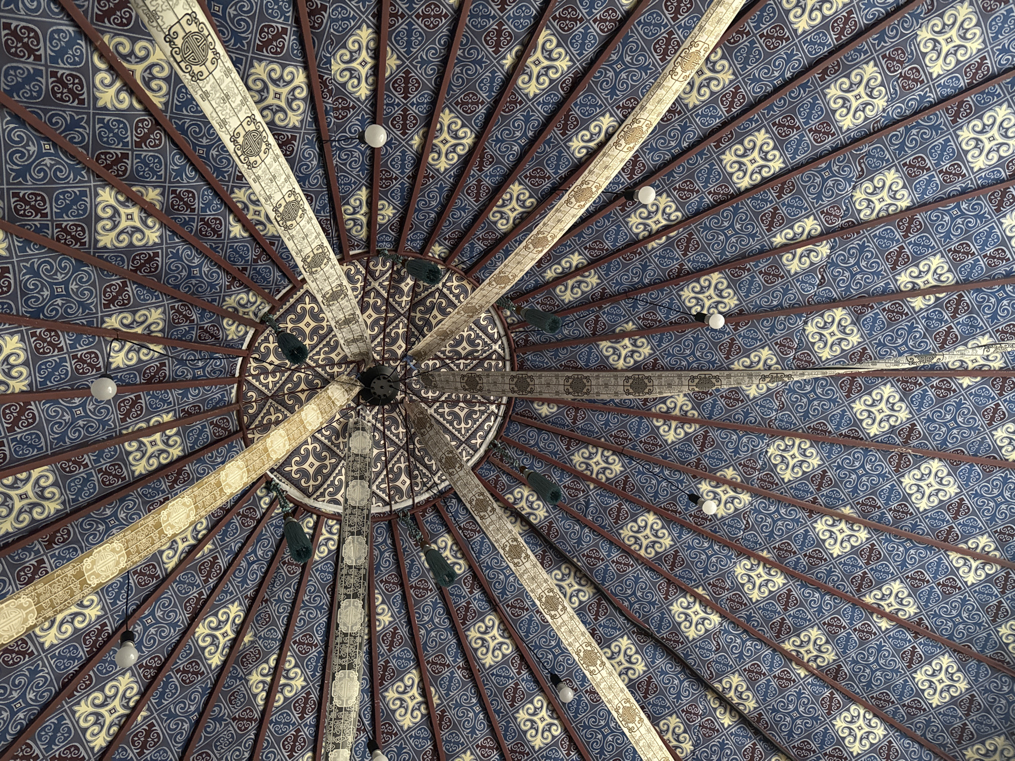 The image shows a close-up, upward view of the intricate ceiling of a traditional yurt in Leskhoz, Kazakhstan. The ceiling is a circular structure with radiating wooden beams, each covered with fabric. The fabric features a complex geometric pattern in shades of blue, yellow, and burgundy, with repeating motifs that appear to be stylized floral or knotwork designs. Interspersed among the main fabric panels are decorative bands of lighter-colored material with ornate, monochromatic patterns. Several small, white light bulbs are visible, attached to wires that hang from the ceiling. Two dark green tassels are also visible, hanging from some of the decorative bands. The overall impression is one of traditional craftsmanship and cultural richness.