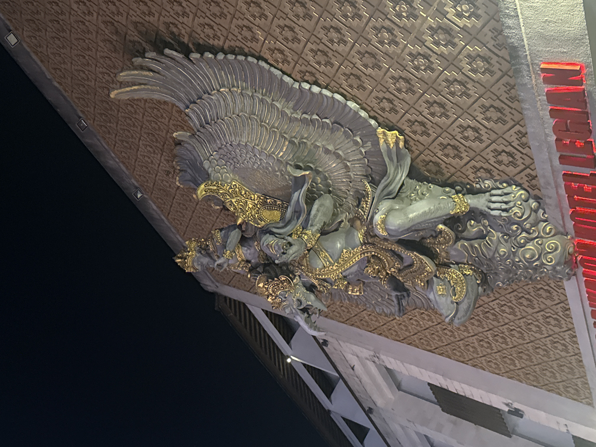 The image shows a large, ornate statue of Garuda, a mythical bird-like creature from Hindu and Buddhist mythology, mounted on the exterior of a building. The statue is predominantly grey with gold accents, displaying detailed feathers, decorative armor, and multiple figures. It appears to be viewed from below and at an angle.
The building itself has a patterned ceiling and modern white structural elements that contrast with the traditional design of the statue. In the background, the night sky is visible. A red sign with the word "NIGHTHAWK" runs down the side of the building.
This particular statue of Garuda is located in Kuta, Bali, Indonesia. It is a landmark often associated with the Garuda Wisnu Kencana Cultural Park but is also commonly found adorning various buildings and gateways throughout Bali.