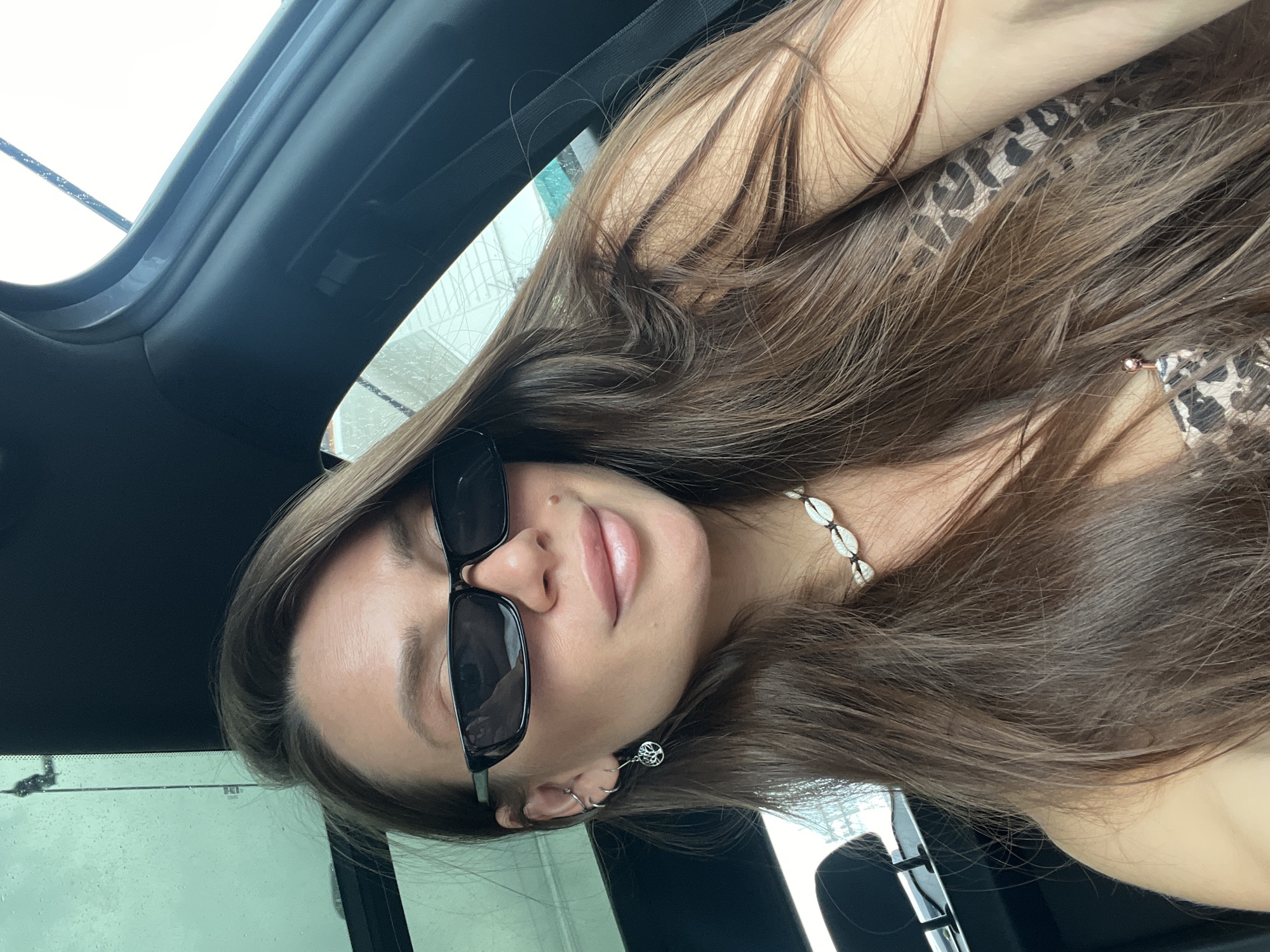 A close-up, upward-angled self-portrait shows a female individual inside a car. Her head is tilted, and she looks directly at the camera with a subtle, closed-mouth smile. She has long, wavy brown hair that falls around her. She wears dark, rectangular sunglasses that obscure her eyes.
Around her neck is a necklace made of white cowrie shells. Her left ear features a small hoop earring and a dangling earring with a 'Tree of Life' design. She is wearing a leopard-print top, visible under her hair.
The setting appears to be a car interior, with parts of the vehicle's dark headliner, A-pillar, and a seatbelt visible. Through the windows, water droplets are seen, suggesting either rain or that the car is in a car wash. The lighting indicates it is daytime. No discernible text is visible.