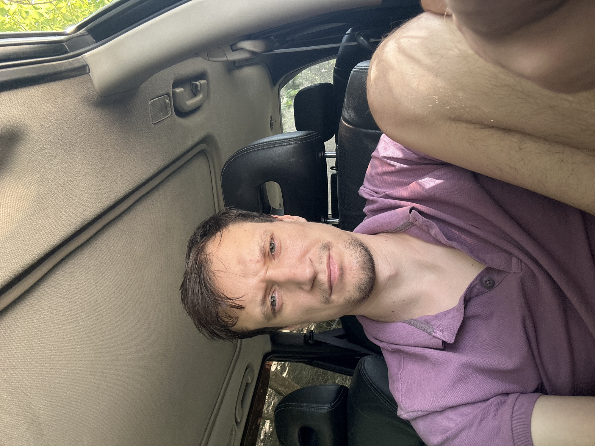 A man is seated in the driver's seat of a car, looking at the camera. He has short brown hair and a light beard. He is wearing a purple polo shirt. The interior of the car is visible, including the headrests of the seats and the car's sunroof. The background shows trees and foliage, indicating an outdoor setting, possibly in the city of Makiv, Ukraine. The man's right leg is propped up.