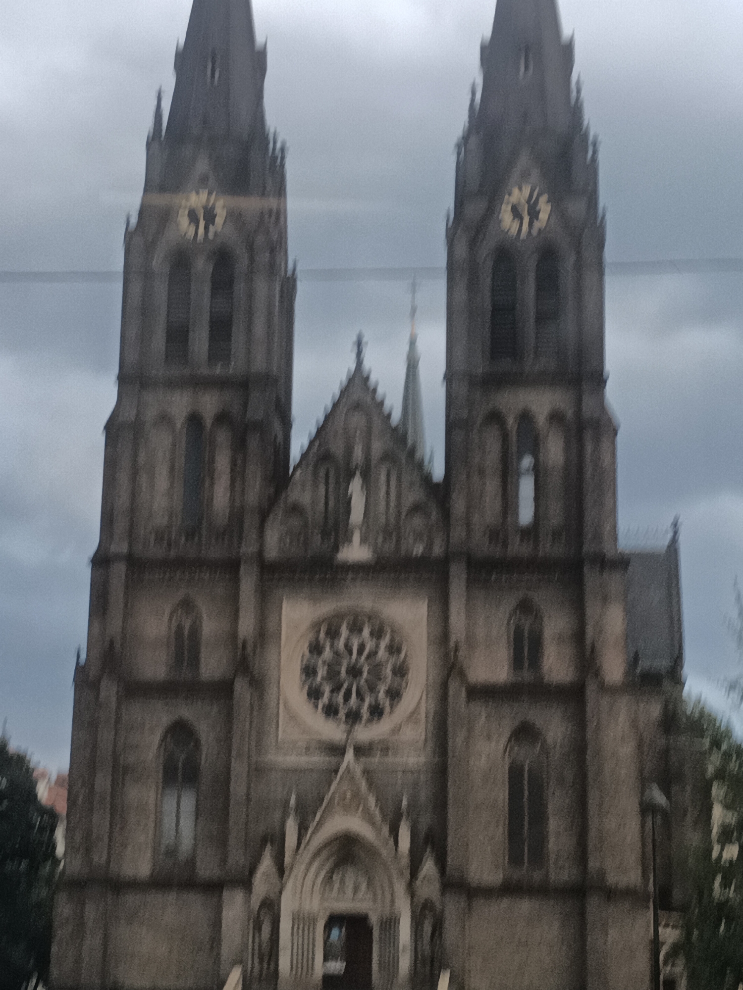 The image shows the exterior of a gothic style church, likely St. Peter and Paul's Cathedral in Vyšehrad, Prague, Czechia. The church has two prominent spires with clocks on them. The sky is overcast. The building appears to be made of stone.