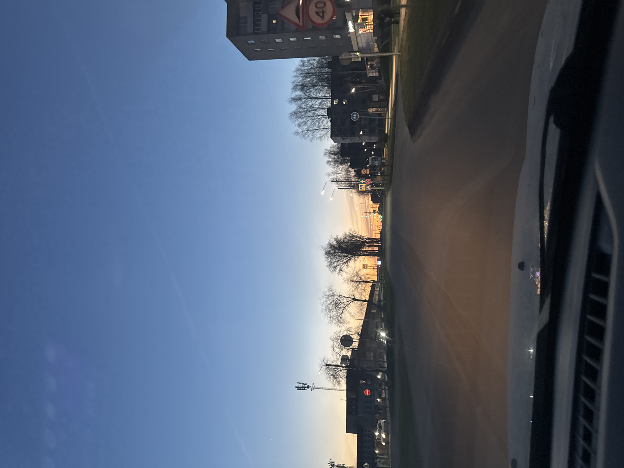 The image presents a view of Chaikovsky, Russia, captured from inside a vehicle. A road stretches into the distance, lined with buildings, streetlights, and bare trees. A building with a "40" speed limit sign is visible. The sky transitions from a deep blue to a lighter hue near the horizon, possibly indicating dawn or dusk. The right side of the image shows part of the vehicle's interior.