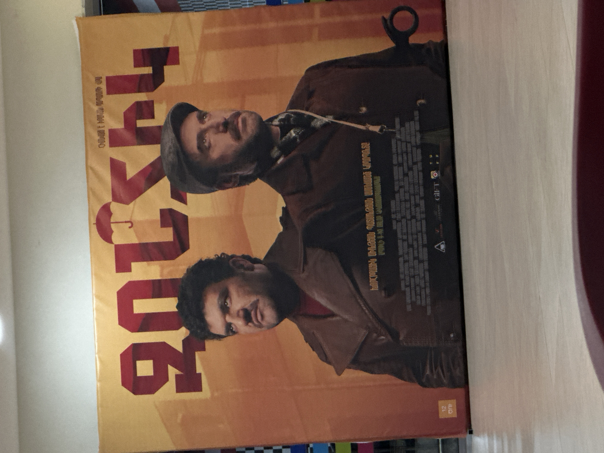 The image shows a movie poster, possibly for the film "206," oriented sideways. The poster features two men, one with a cap and the other with curly hair, both wearing coats. Text in Armenian is present alongside the title. The poster has a yellowish-orange color scheme. The location appears to be indoors, with a table visible in the foreground.