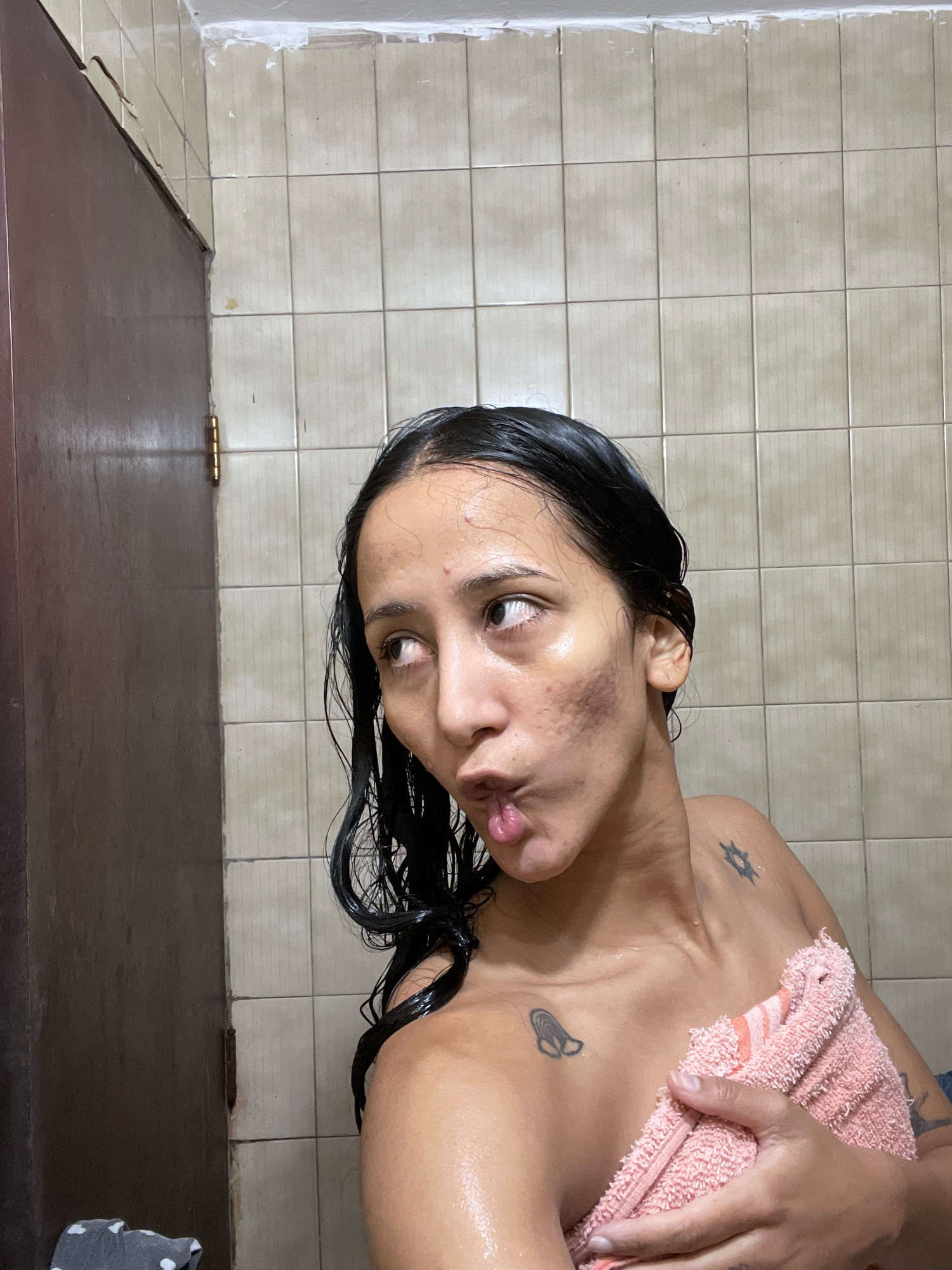 A woman with wet, dark hair is holding a pink towel to her chest, standing in front of a tiled wall. Her skin is glistening with water. She is looking to her left with her lips pursed. There are tattoos on her shoulder and collarbone. To her left is a brown door with visible hinges. The room appears to be a bathroom, likely in a residential building. This image does not contain enough information to determine its location in Porlamar, Venezuela.