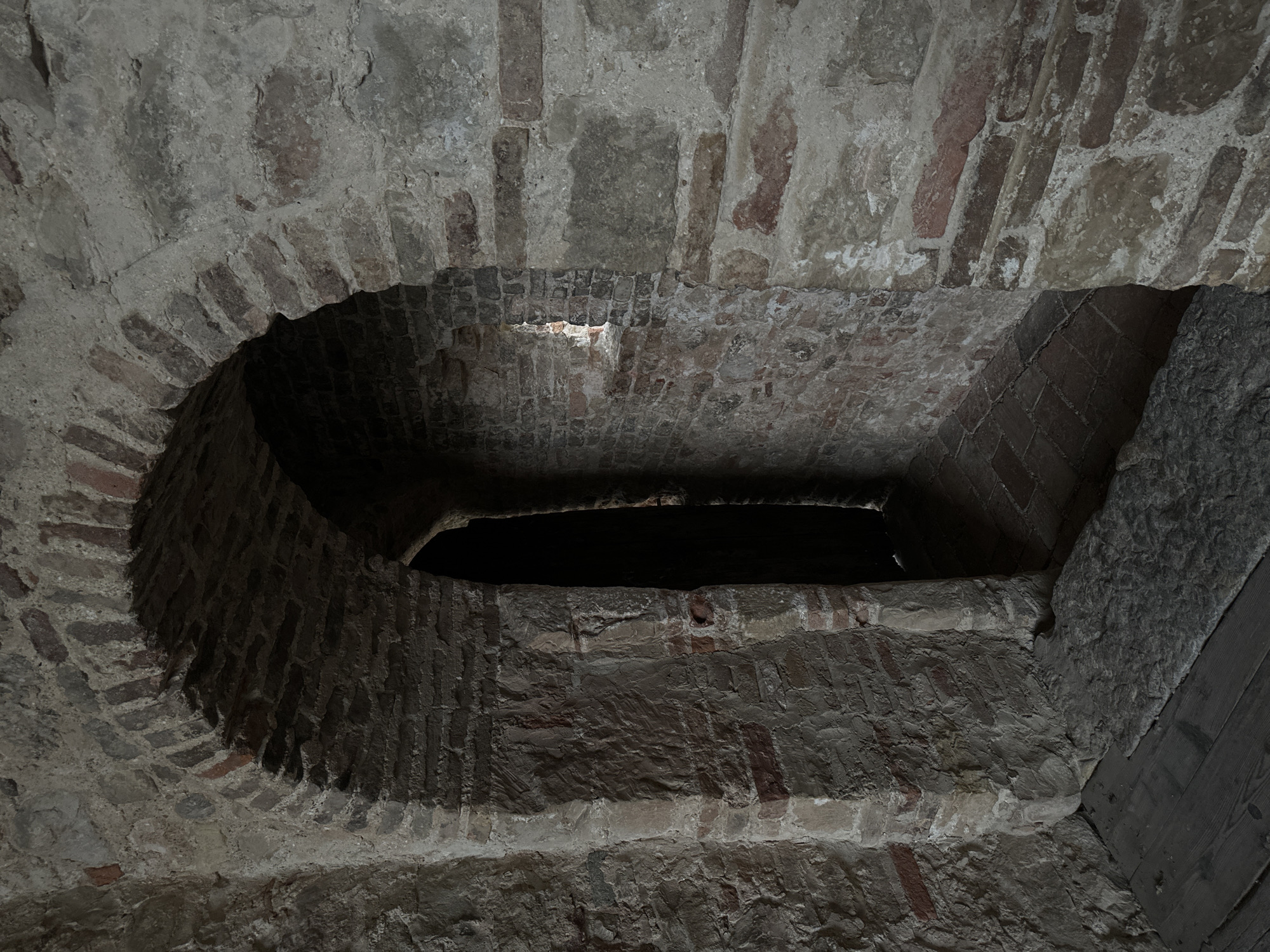 This is a photograph of an arched stone doorway leading into a dark passageway, likely within a historical building in Maser, Italy. The doorway and surrounding walls are constructed from a combination of rough-hewn stones and red brick, with visible mortar. The archway is well-defined, with a series of bricks forming its curve. To the right of the doorway, a small, narrow opening or window is visible in the brick wall. The floor in the foreground consists of wooden planks, with a large, flat stone placed over them near the entrance. The passageway beyond the arch is cast in deep shadow, with only hints of a brick floor visible. The overall impression is one of age and solidity, with a sense of mystery emanating from the dark interior.
