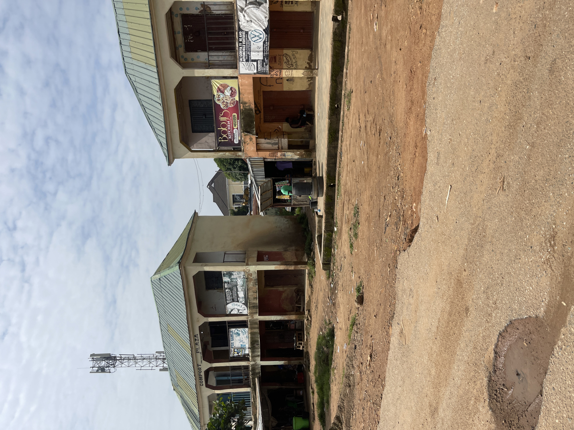 The media file captures a street view in Gwong, Nigeria, on a cloudy day. The scene is dominated by two adjacent buildings, likely shops or small businesses, with two stories each. The buildings have a somewhat weathered appearance, with signs of age and some peeling paint.
The lower building, on the left, displays several signs, including "COMFORT PLAZA," "AN NAMIJI," and "BABZEE." Further down, another sign reads "Babyji's KITCHEN." A prominent sign on the upper level of this building advertises "SAI GODIYA TY RE AND AUTOMOBILE VENTURES," featuring a Volkswagen logo. The shops on the ground floor appear to be open, with some wares visible inside. A person is seated in one of the shops.
The building to the right has a similar two-story structure, with barred windows and shopfronts. One shop has a large banner with pictures of food items. Further up, another banner with a silhouette of people is visible. The entrances to these shops are open, revealing dimly lit interiors.
A dirt road runs alongside the buildings, with a section of paved road visible to the right, showing a large puddle of muddy water. Beyond the buildings, a telecommunications tower is visible against the sky, indicating the presence of infrastructure in the area. The overall impression is of a typical bustling commercial street in a Nigerian town, with a mix of services and shops. The lighting suggests it is daytime.