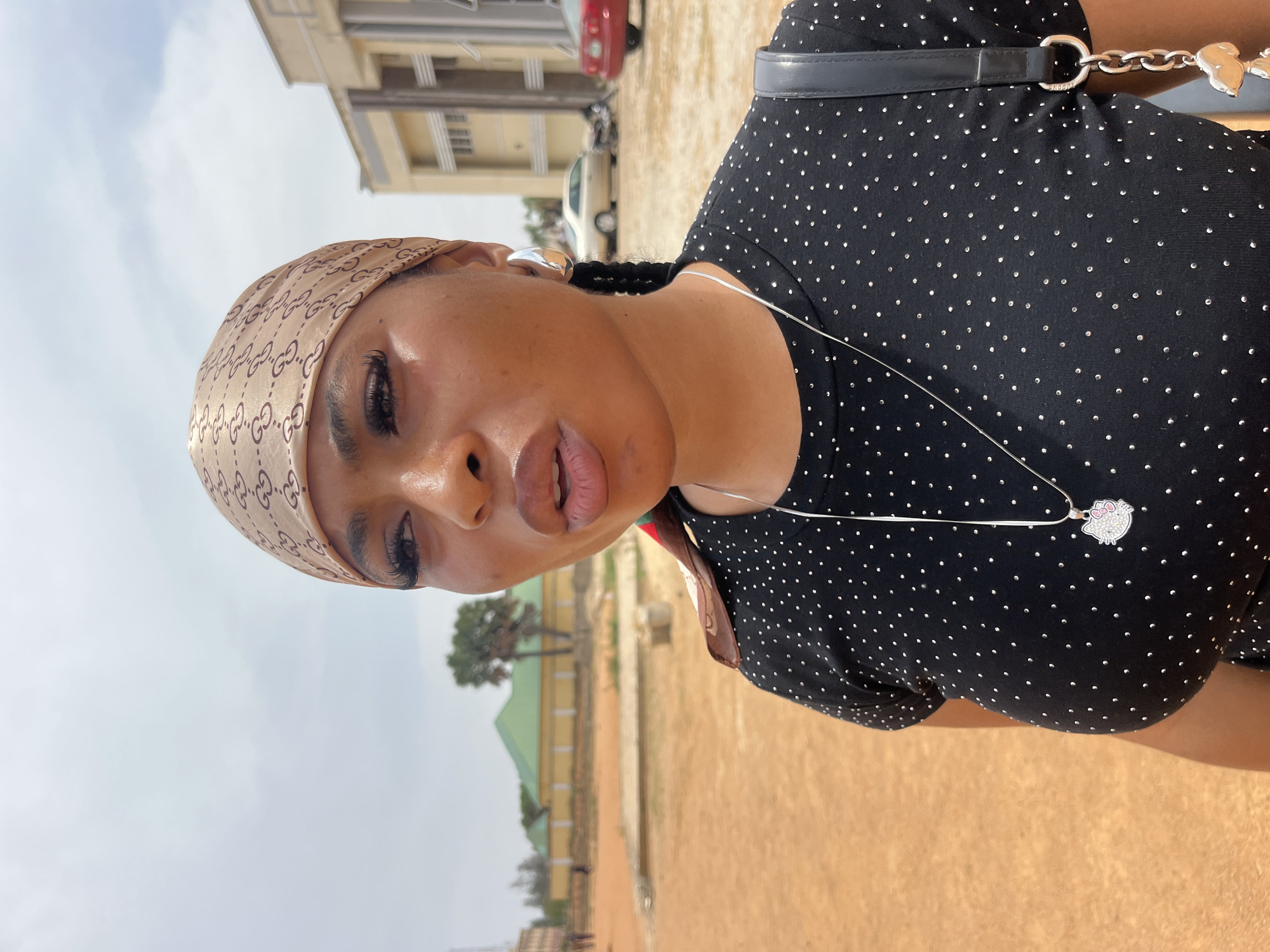 A young woman is the central subject of this outdoor photograph, captured in a half-body shot with her head slightly tilted and her gaze directed towards the viewer. She wears a light brown headscarf adorned with a repeating "GG" pattern, indicative of the Gucci brand. Her visible ear features a large, silver-toned earring, and her dark hair is styled in braids peeking out from under the scarf. She is wearing a black top or dress embellished with numerous small, sparkling silver studs or rhinestones. A thin silver-colored necklace with a distinct white pendant, shaped like Hello Kitty with a pink bow, hangs around her neck. A black strap, likely from a bag, is visible over her shoulder.
The setting appears to be an open outdoor area under a bright, cloudy sky, suggesting daytime with soft lighting. In the background, there are various buildings, including one with a light cream or beige facade and another with a green roof. Several cars are parked in the distance. The ground is an unpaved, light brown or reddish-brown dirt or sandy surface. The woman's expression is neutral to slightly pouty, indicating she is posing for the camera.