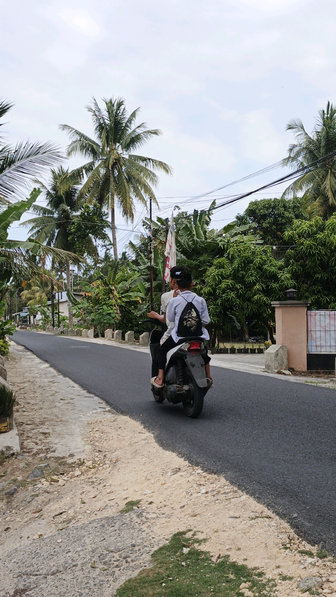 Two individuals, one driving and one riding as a passenger, are on a scooter on an asphalt road in a tropical environment. The driver, wearing a dark cap and a light-colored shirt, is partially obscured by the passenger. The passenger, in a white shirt and dark pants, carries a black backpack with a white design. They are holding a flag on a pole that extends above the passenger's head. The road is flanked by a dirt and gravel shoulder on the left and a paved walkway. Lush green vegetation, including palm trees and banana trees, lines the roadside. In the background, there are houses and a stone pillar with a fence. The sky is overcast with some visible clouds. The scene appears to be in Cintaratu, Indonesia, suggested by the context.