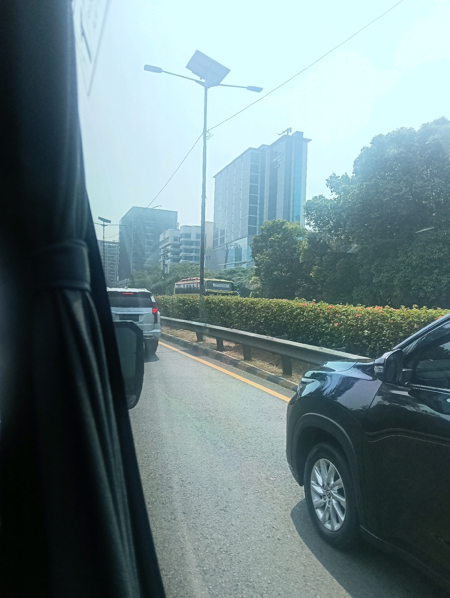 The image depicts a street scene in East Jakarta, Indonesia, likely during daylight hours. The perspective is from inside a vehicle, with a dark fabric covering the left side of the frame, possibly a curtain or a portion of the vehicle's interior.
On the road, a white SUV is visible in the middle lane, its rear facing the camera. To the right of the SUV, a dark-colored car is in the foreground, its front section and wheel clearly visible. Both vehicles appear to be in traffic.
The road has distinct yellow and white lane markings. A metal guardrail separates the traffic lanes from a landscaped area. This area features lush green bushes and some flowering plants with orange-pink blossoms. Beyond the greenery, several modern buildings of varying heights are visible, suggesting an urban environment. One prominent building is a tall, multi-story structure with a glass facade. In the distance, a yellow bus is partially visible.
Overhead, a street light with a solar panel is mounted on a pole. The sky is hazy or overcast, contributing to a diffused light. The overall impression is a busy urban street with moderate vehicular traffic and modern infrastructure.