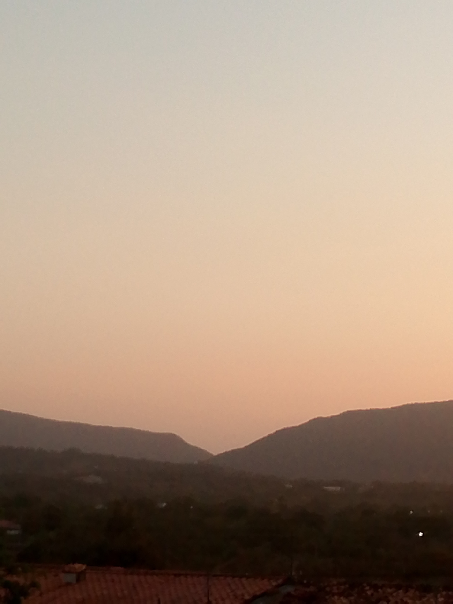 The image captures a view of mountains and vegetation near Juan Griego, Venezuela, during a sunset. The sky is a gradient of orange and light tan, indicating twilight. The mountains are dark silhouettes against the bright sky. In the foreground, there are rooftops suggesting buildings in the area. The overall scene evokes a quiet, natural setting within the urban landscape of Juan Griego.