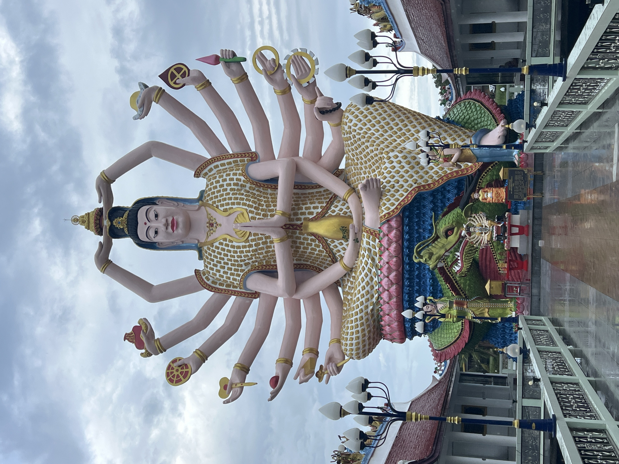 This is a medium shot of a large, colorful statue of a multi-armed deity in front of a building with ornate architectural details and a cloudy sky. The statue, predominantly pink and gold, depicts a serene figure with many arms, each holding a different symbolic object such as a lotus, a wheel, or a small scepter. The deity is adorned with a patterned robe and elaborate jewelry. Behind the main statue, a vibrant green dragon sculpture is visible, with blue and pink decorative elements. The building in the background features red-tiled roofs and white columns. Two lampposts with multiple white lights stand on either side of the scene. The overall impression is one of religious or cultural significance, likely in a temple or shrine setting. The cloudy sky suggests it might be late afternoon or an overcast day. There is a small sign with text in front of the dragon sculpture, but the text is not legible. The location is identified as Baan Plai Laem, Thailand.